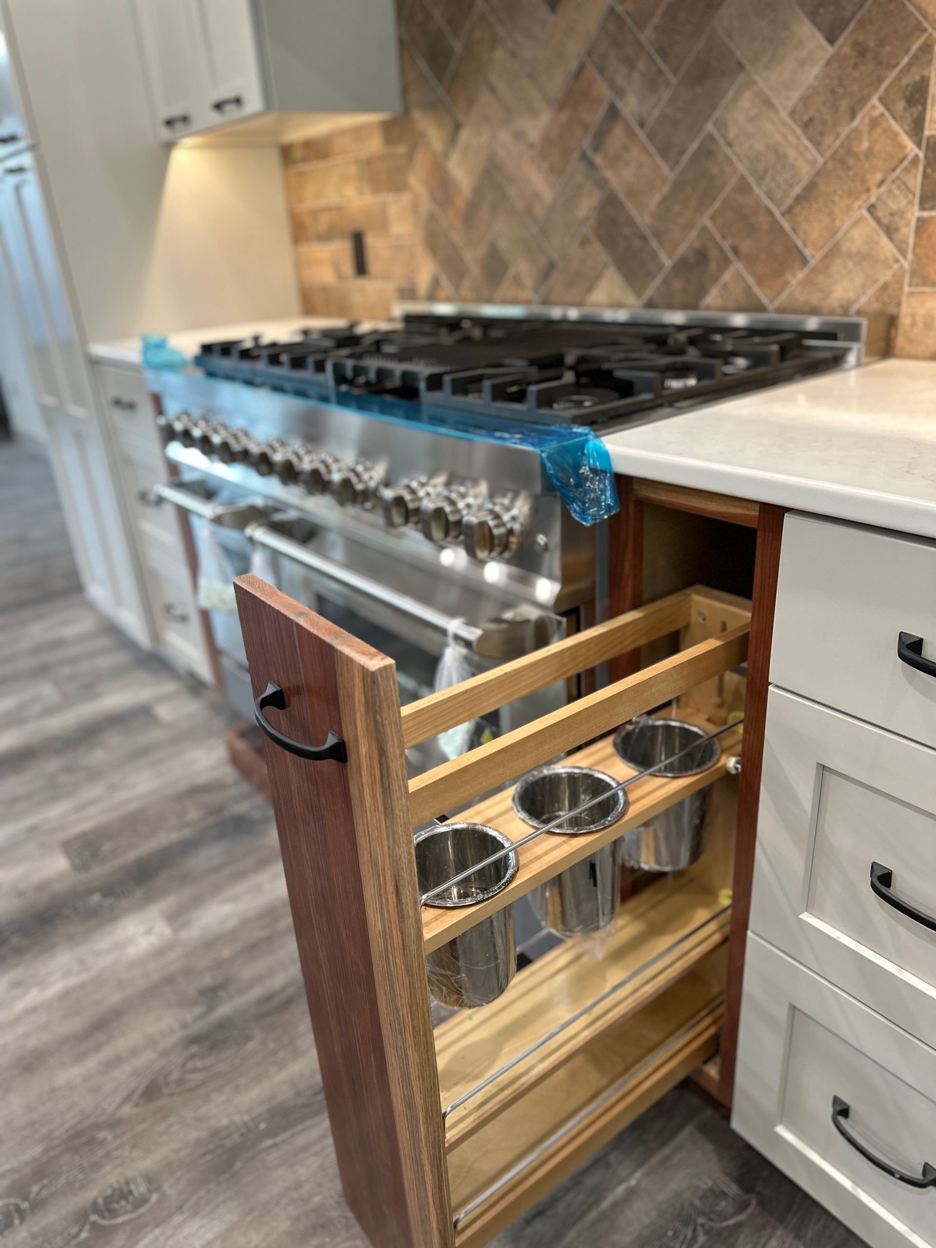 Pull-out spice rack next to a large stainless steel range. Kitchen has white cabinets, wood accents, and a brick backsplash.