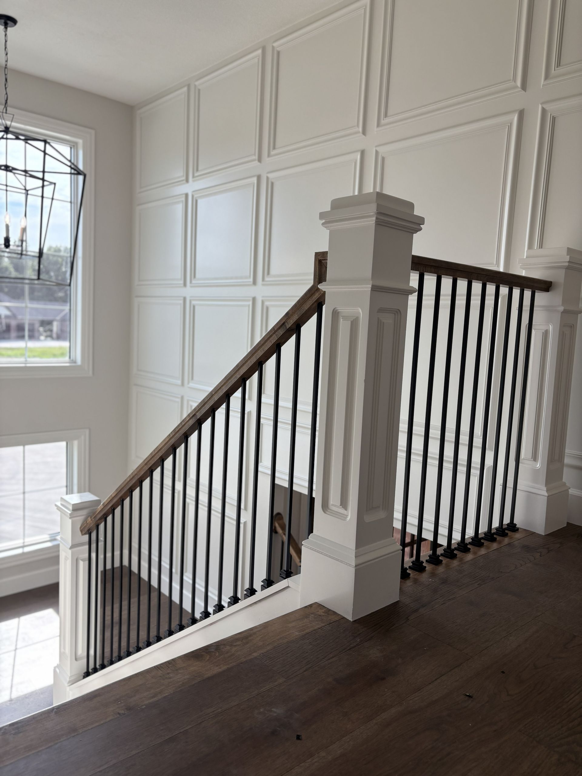 Staircase with wooden handrail and black iron balusters, white paneled wall, and large window.