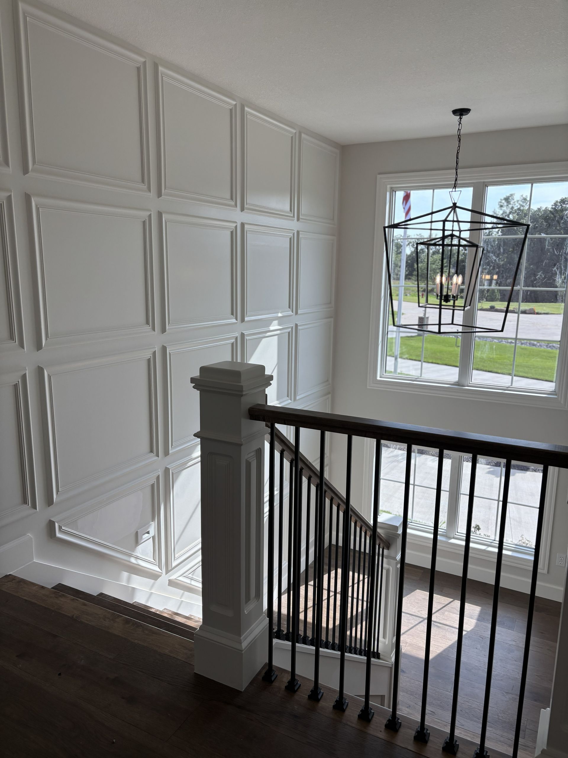 Staircase with white paneled wall, black railing, and modern chandelier, sunlight streaming through a large window.