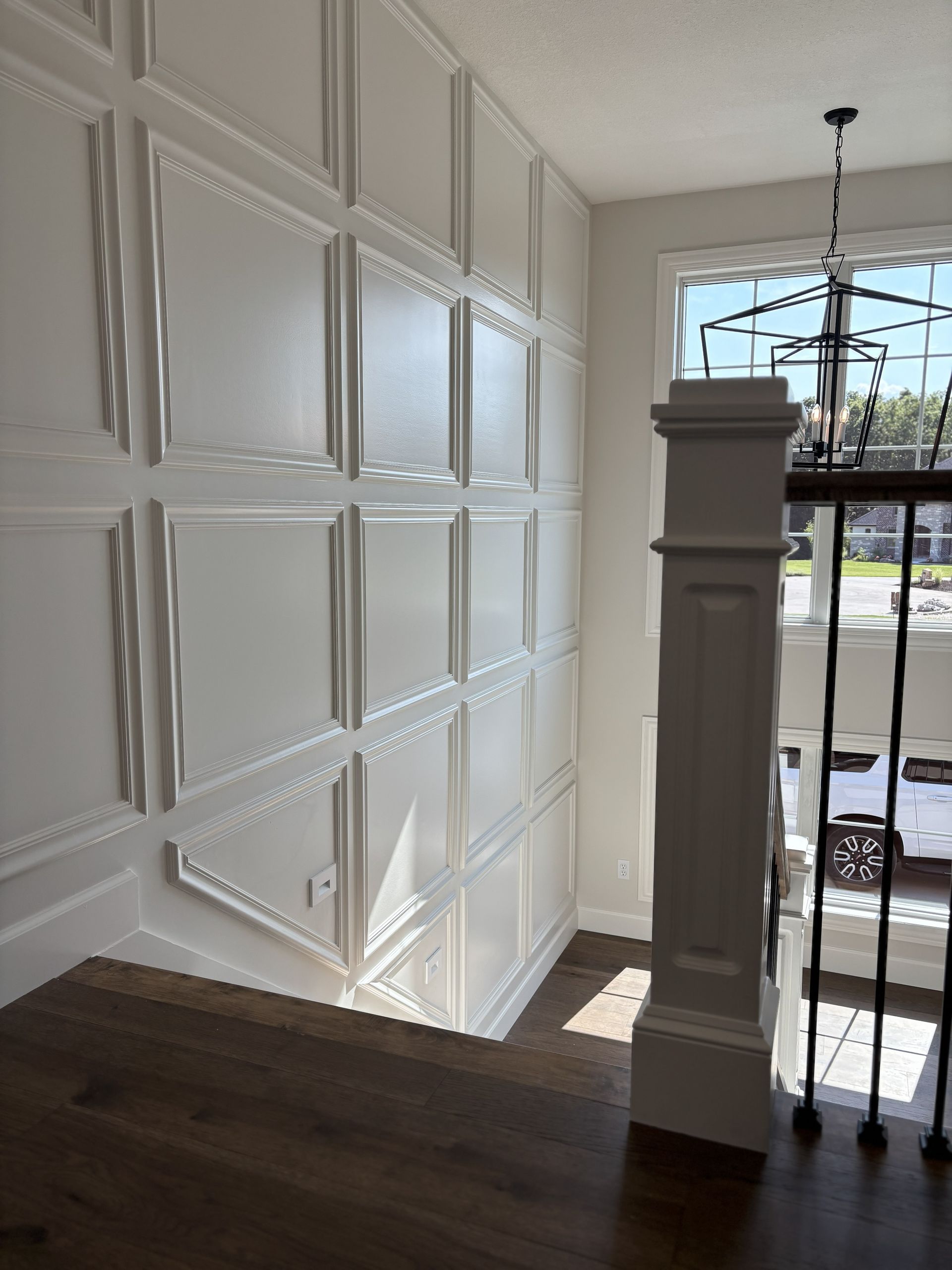 White paneled wall in stairwell with wooden floor and railing. Sunlight through window.