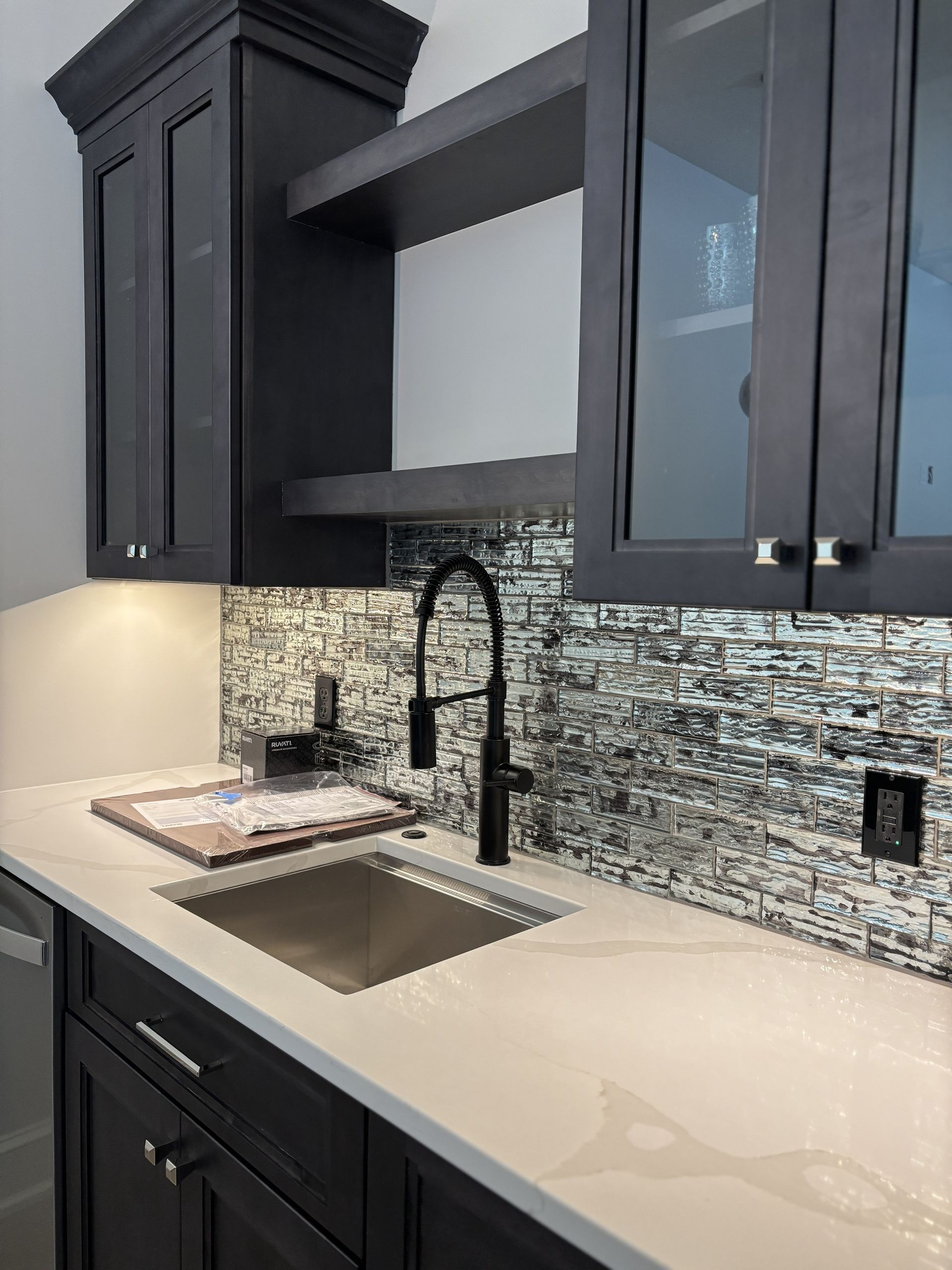 Dark-toned kitchen sink with black faucet, light countertop, and mosaic backsplash, under cabinets.