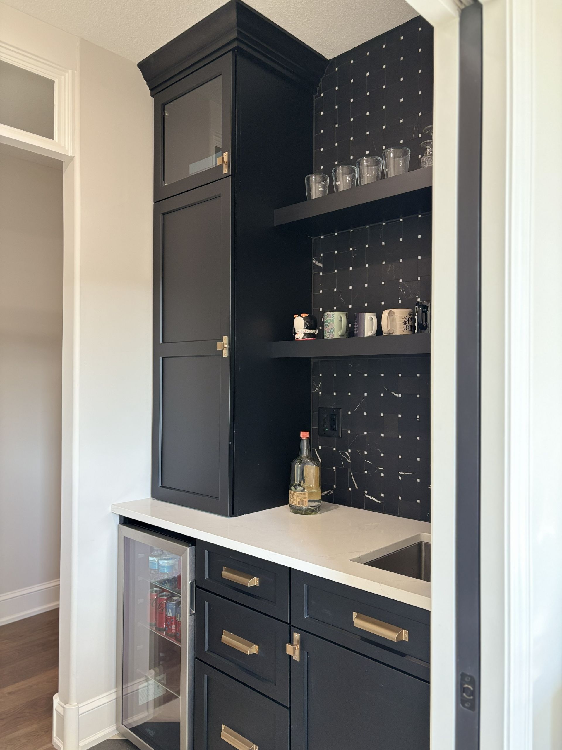 Black cabinet with countertop, mini-fridge, and sink. Shelves hold glasses and mugs against a patterned backsplash.
