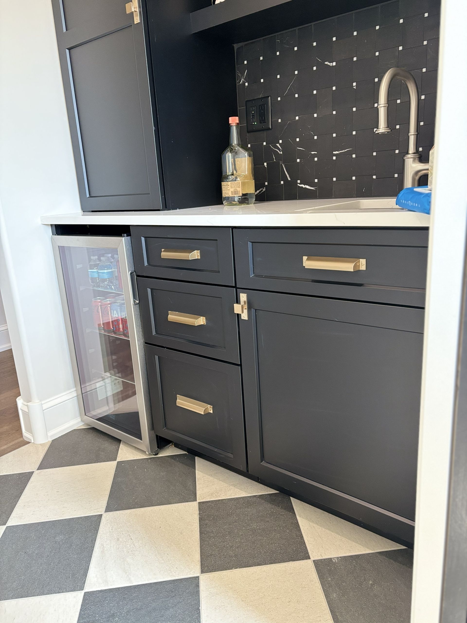 Black cabinets with gold hardware, mini-fridge, and sink. White countertop, checkered floor.
