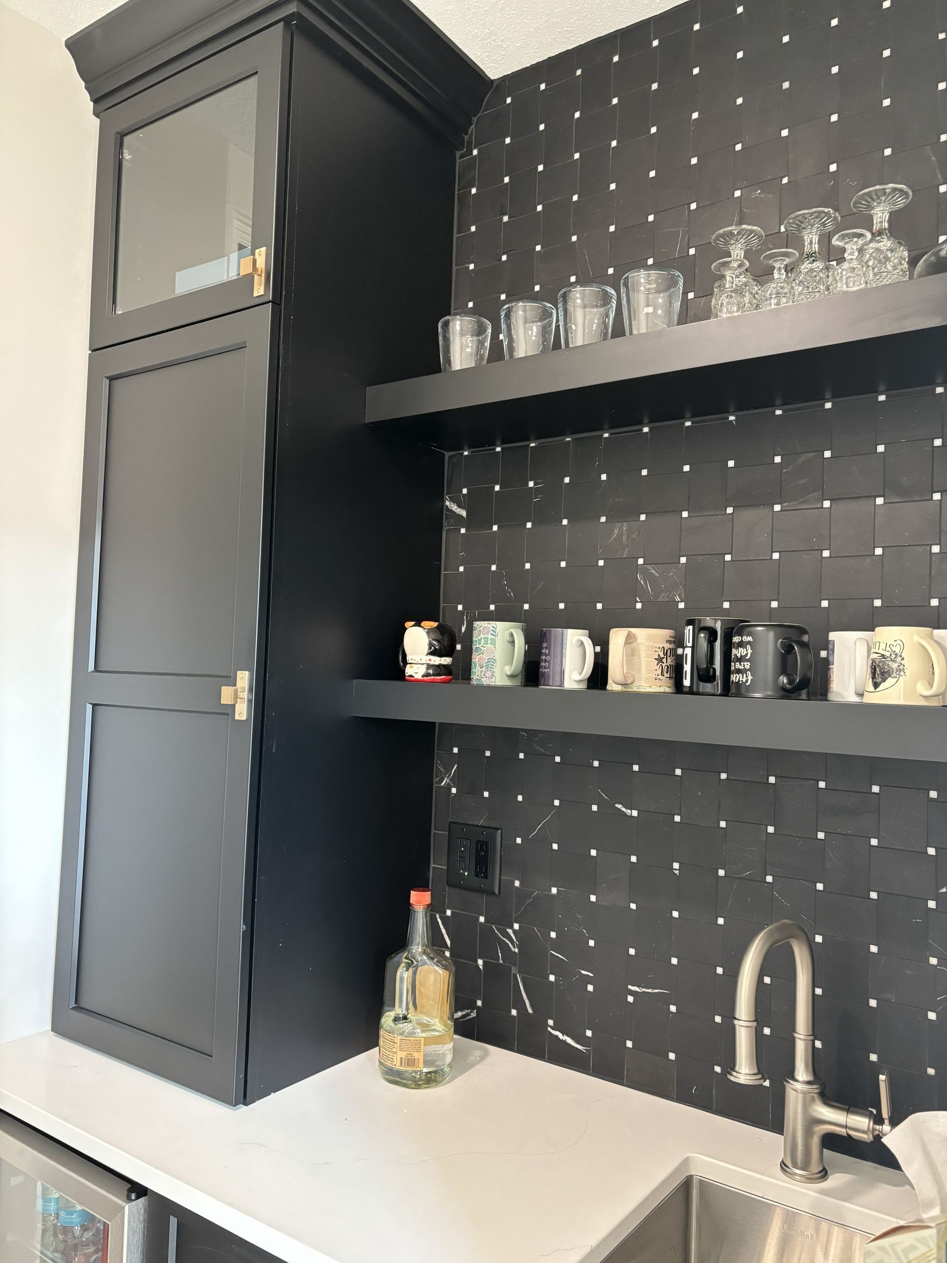 Black bar cabinet and shelves with mugs and glasses. White countertop and black tile wall.