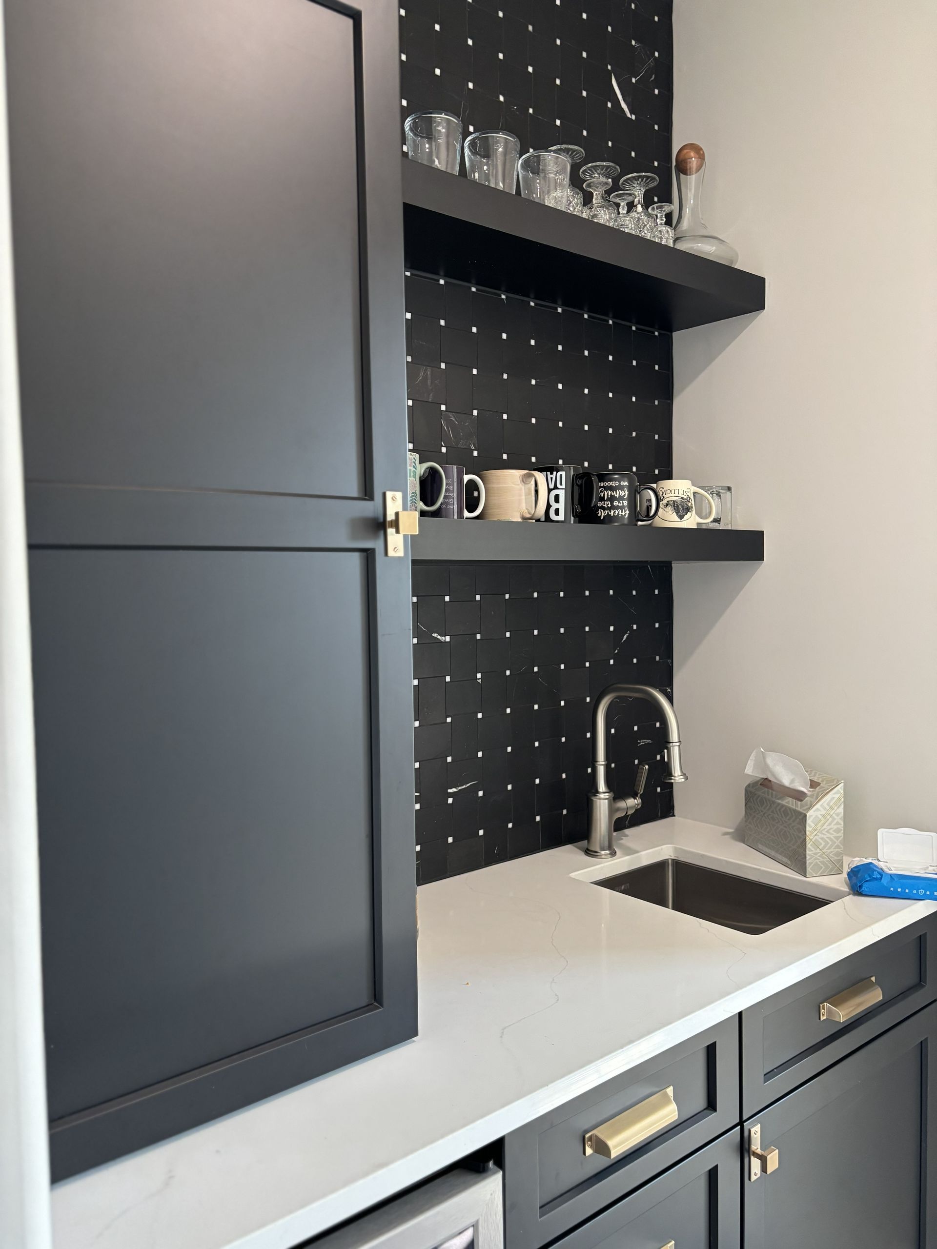 Black wet bar with sink, shelves, and cabinets. A black pegboard backdrop is behind the shelves, which display mugs and glassware.