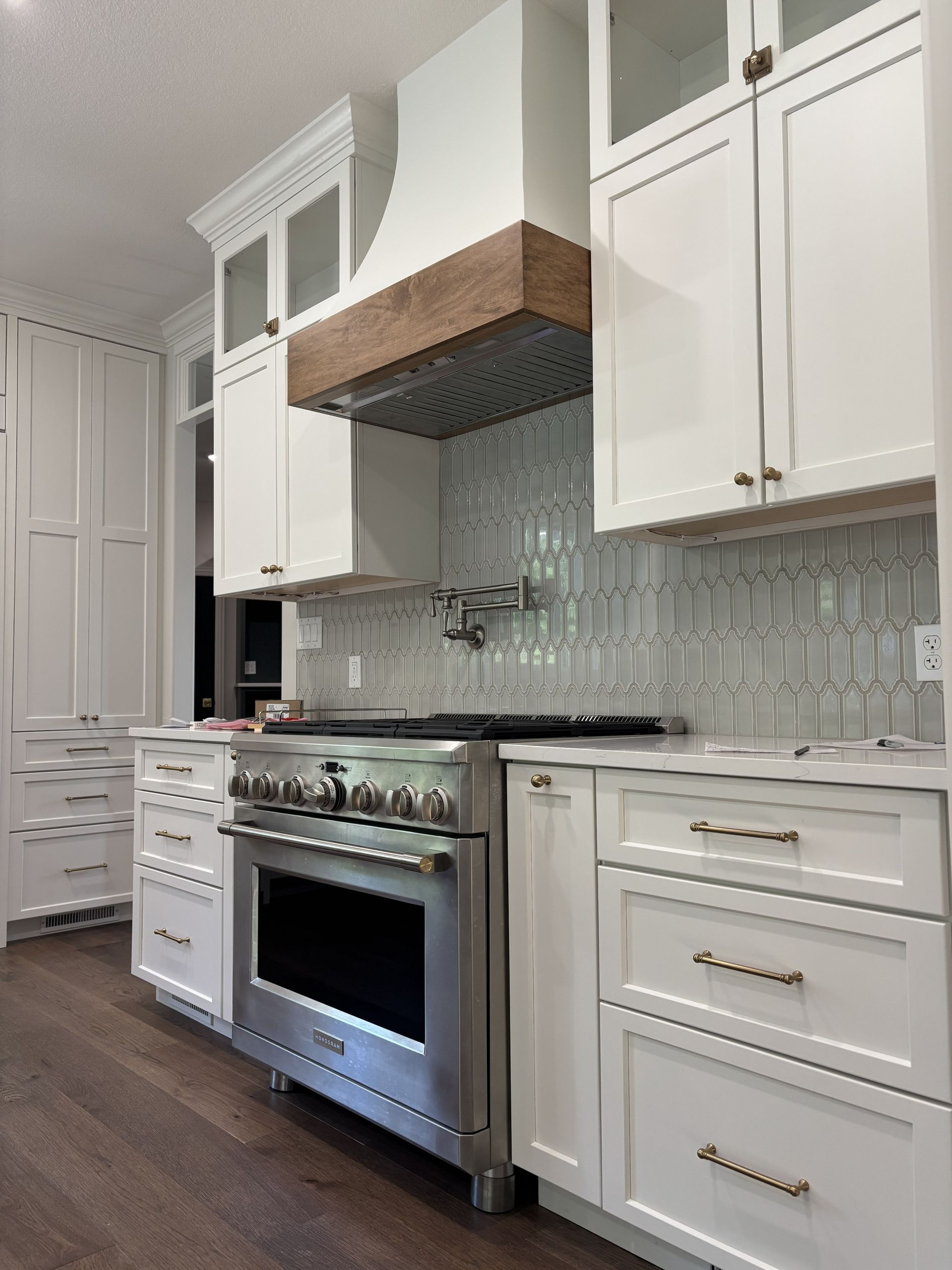 White kitchen with stainless steel oven, wood range hood, and light blue tiled backsplash.