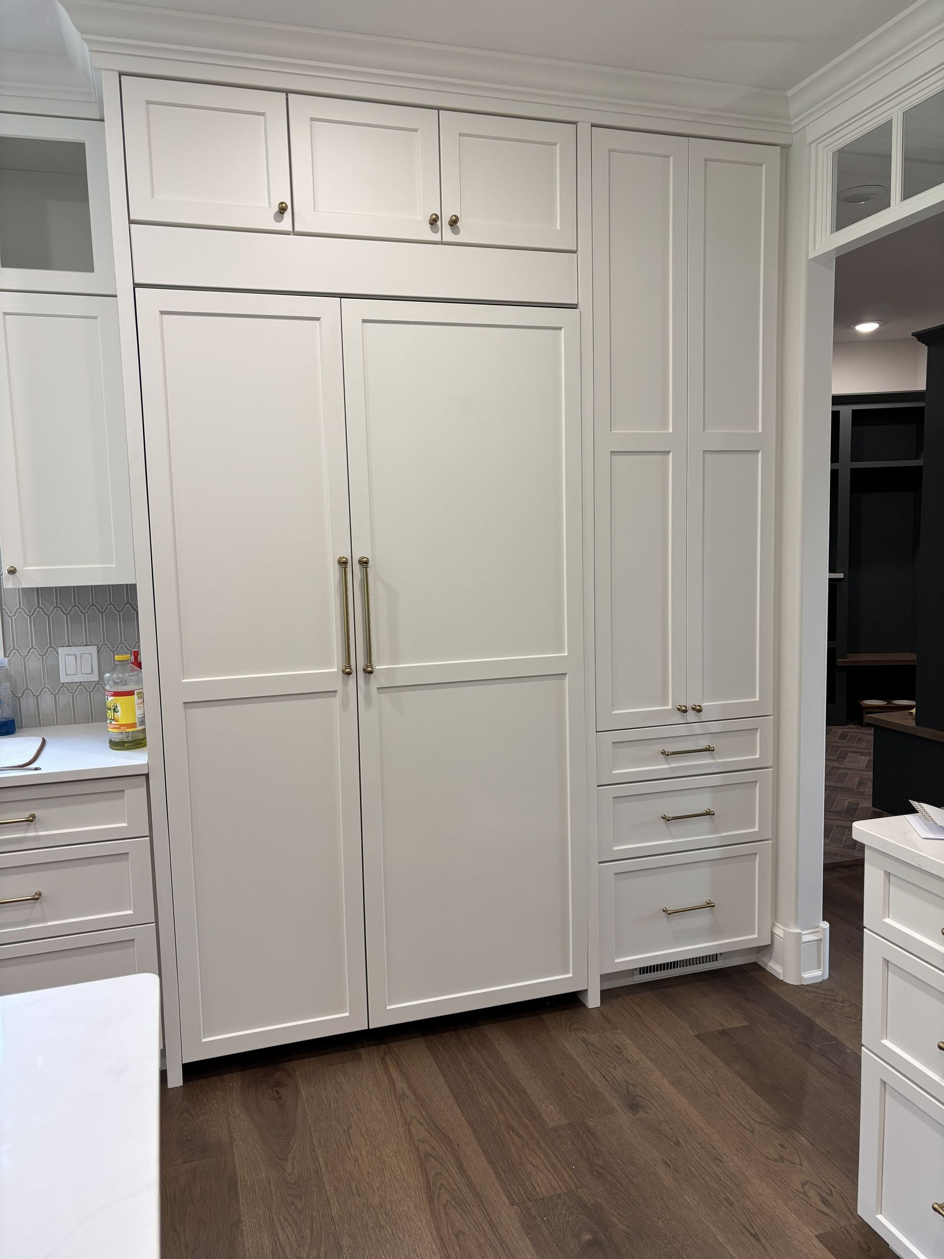 White built-in kitchen cabinets with refrigerator, drawers, and upper storage against a wall with hardwood flooring.