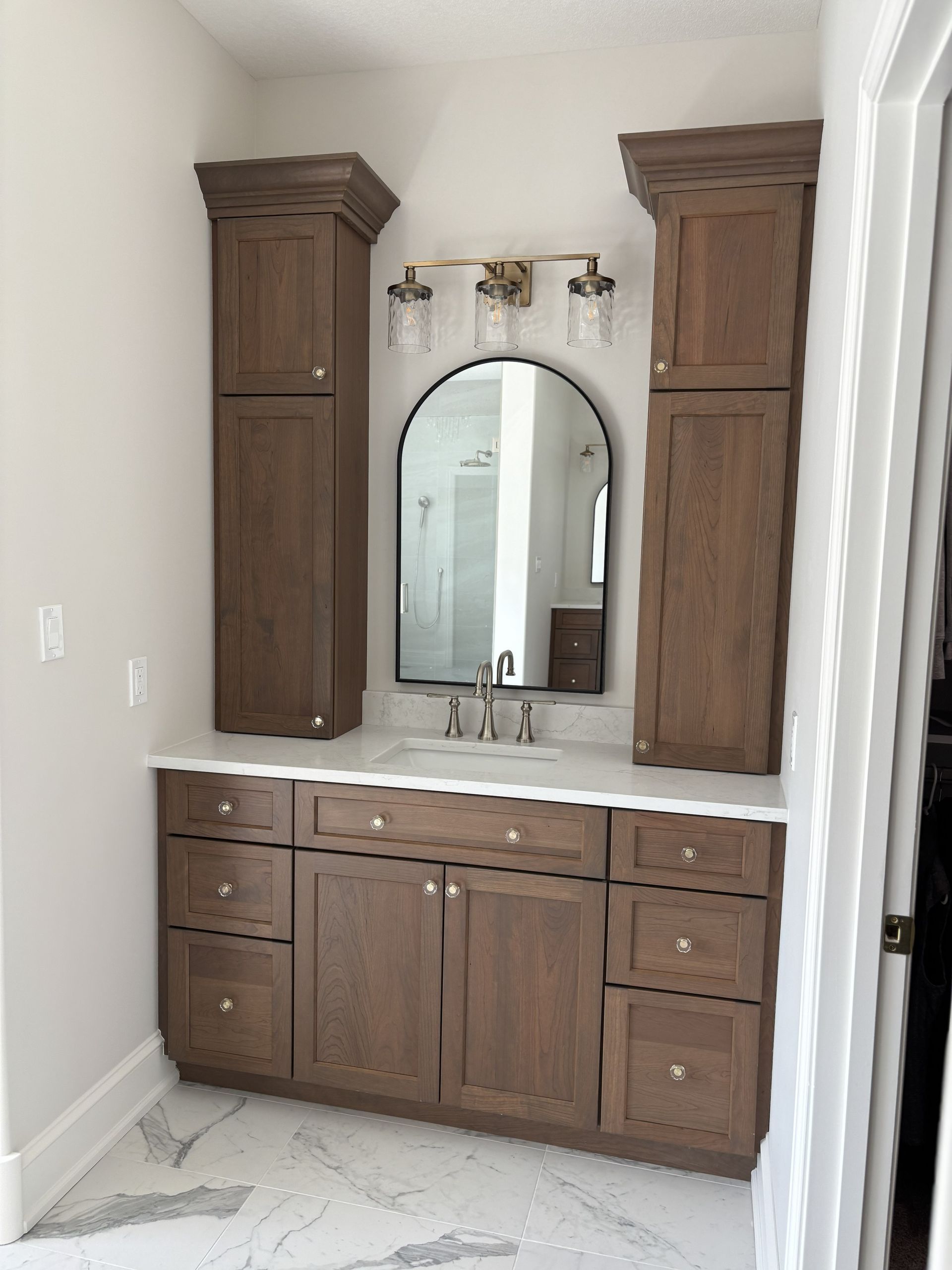 Bathroom vanity with dark brown cabinets, white countertop, arched mirror, and gold fixtures.