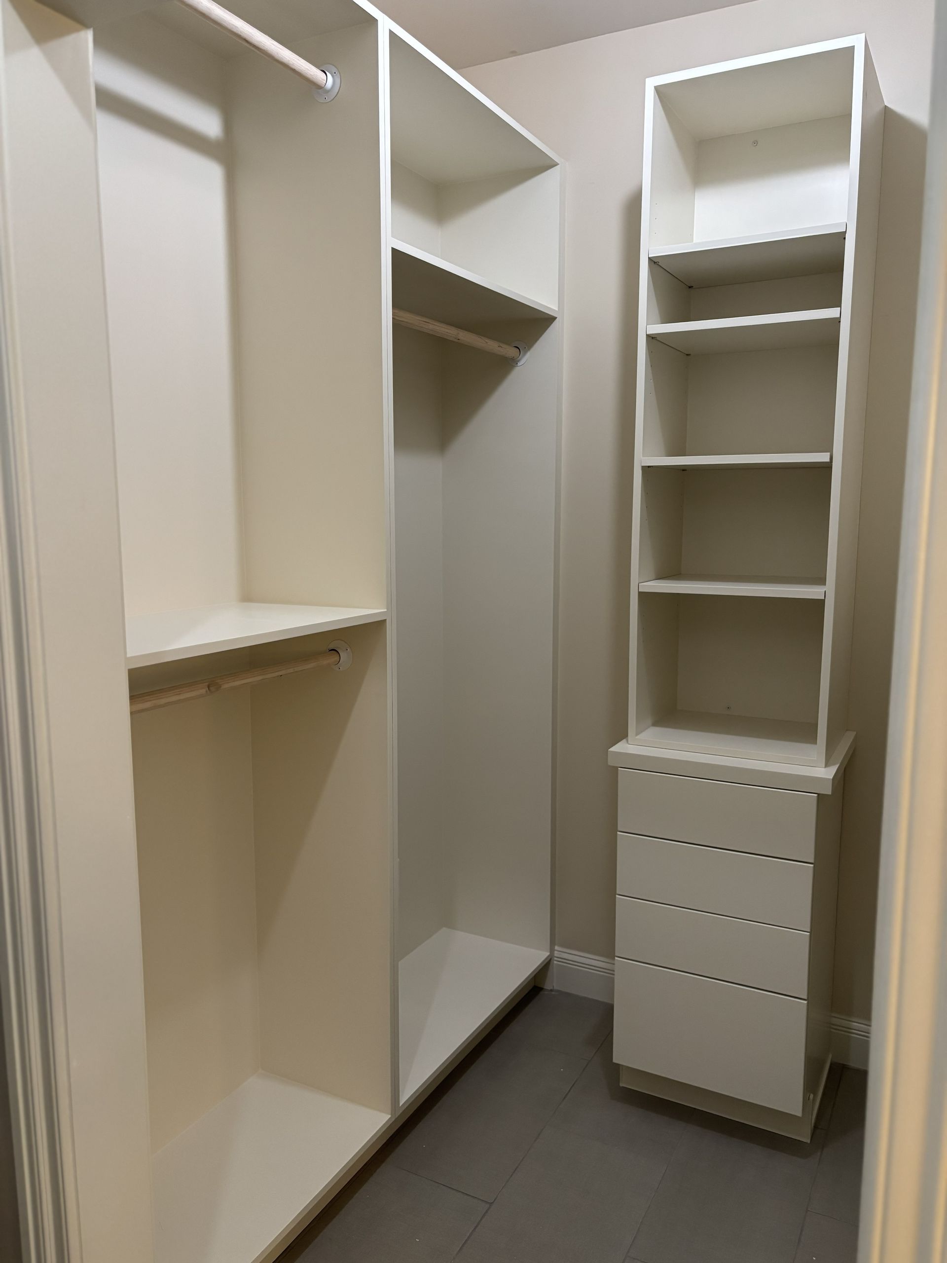 Walk-in closet with white shelves, drawers, and hanging rods. Cream-colored walls, gray flooring.