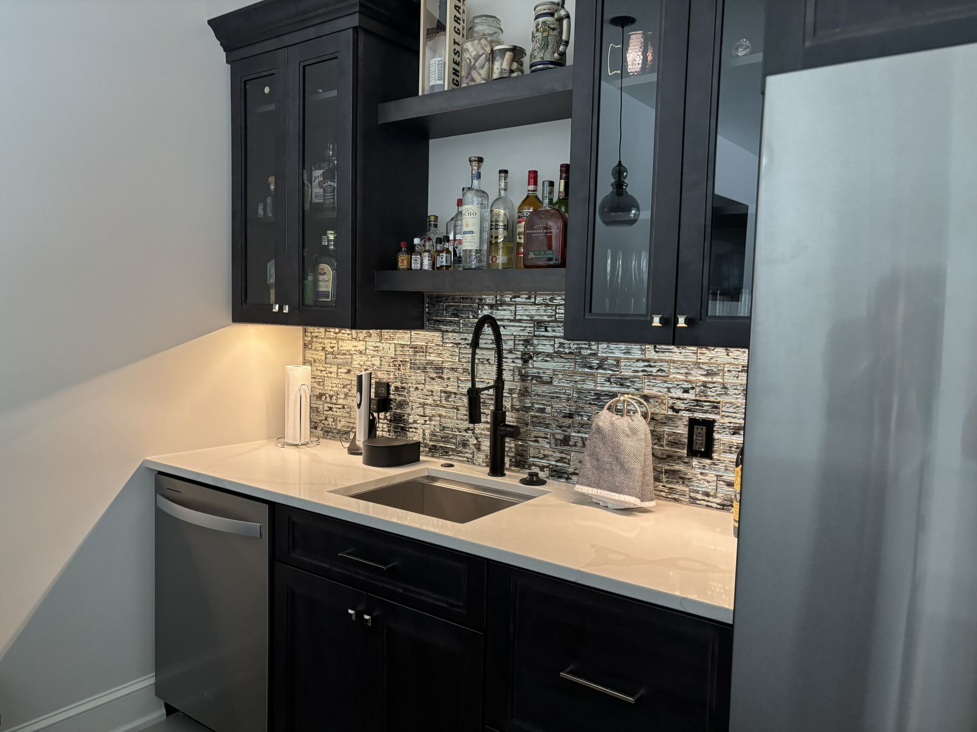 Black bar cabinet with sink, liquor bottles, and backsplash tile against a white countertop.