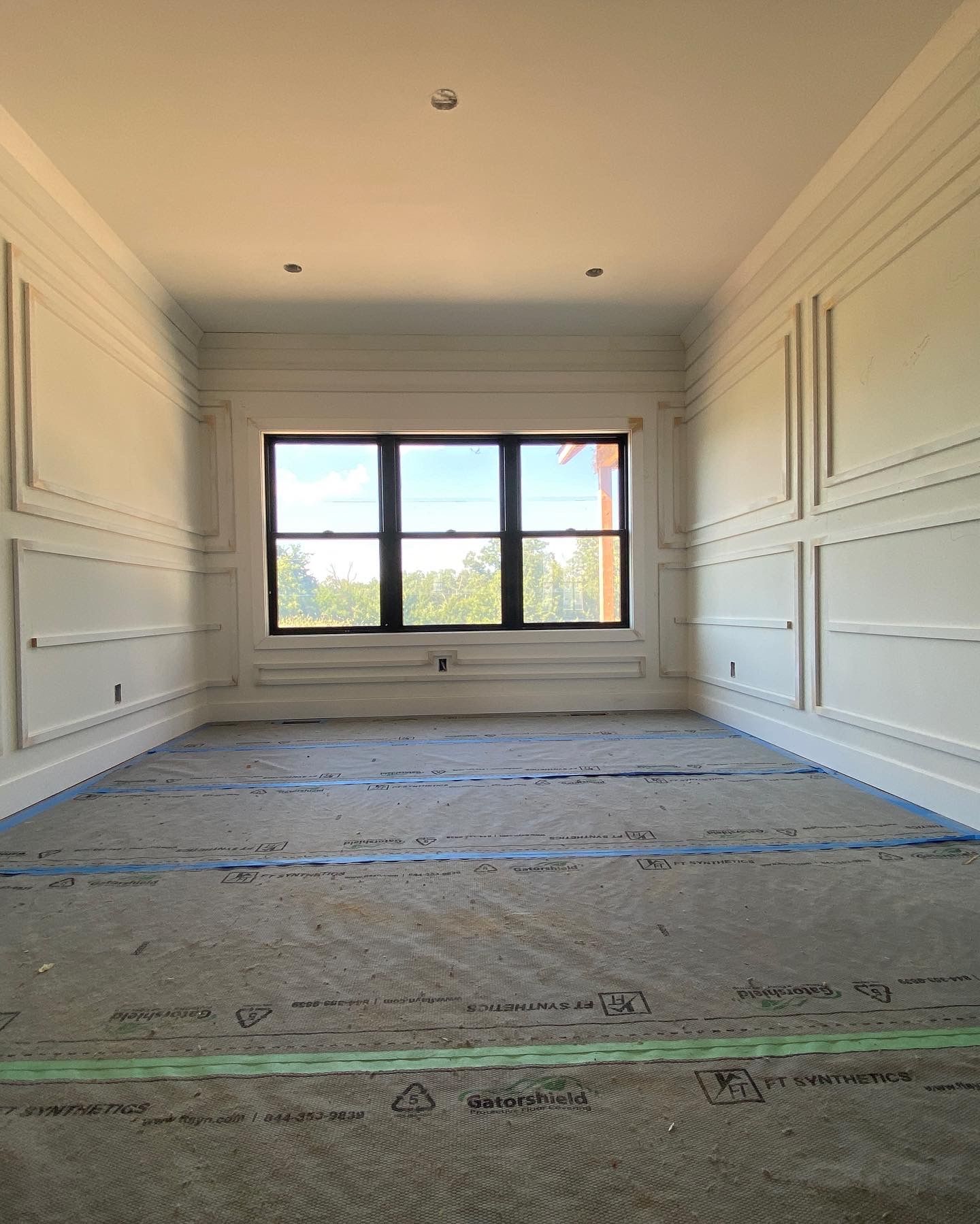 Empty room with white wall paneling, black window, unfinished floor.