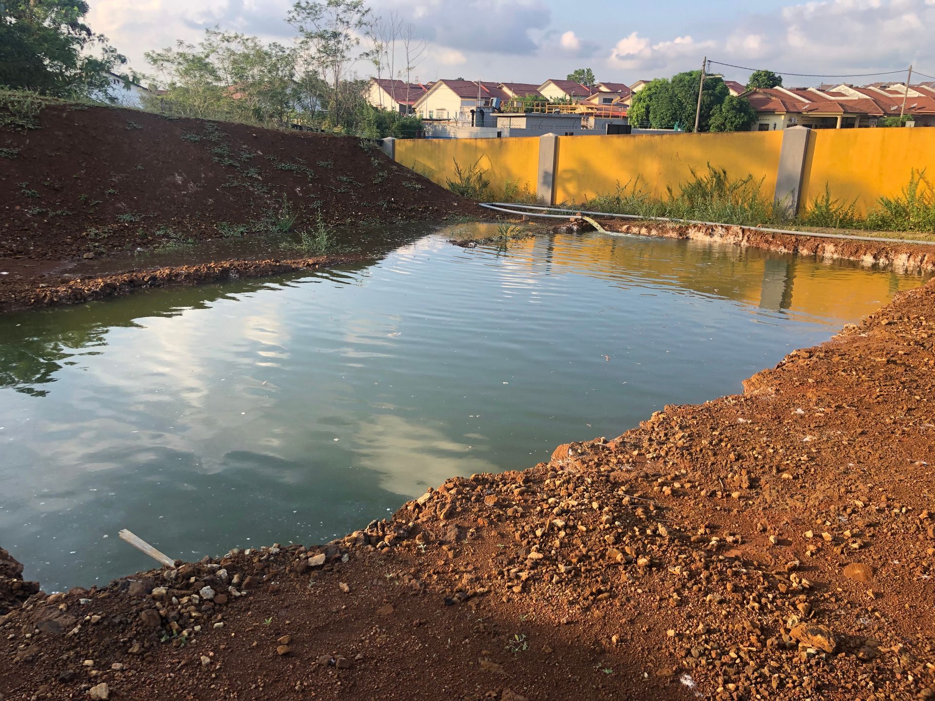 Large muddy pond with gravel banks, a yellow fence, and houses in the background under a cloudy sky
