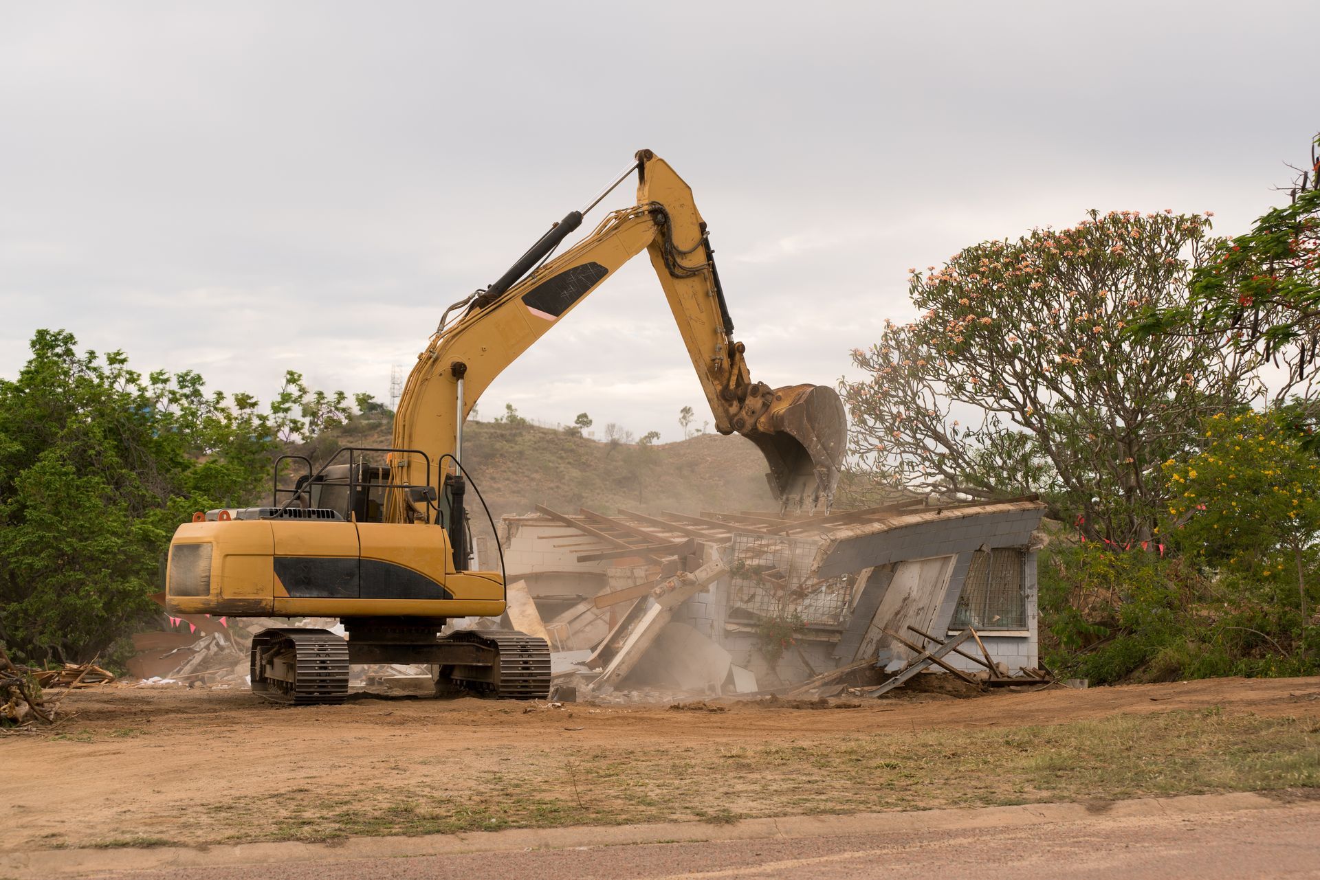 Yellow excavator demolishing a small building amid dust and debris, surrounded by trees.