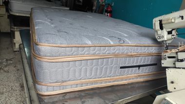 Colchón gris sobre un somier dentro de un taller, con piezas de una máquina de coser visibles en el borde.