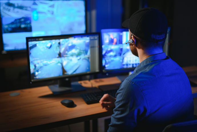 Man monitoring multiple commercial security camera screens in a dim room, wearing a cap and earpiece.