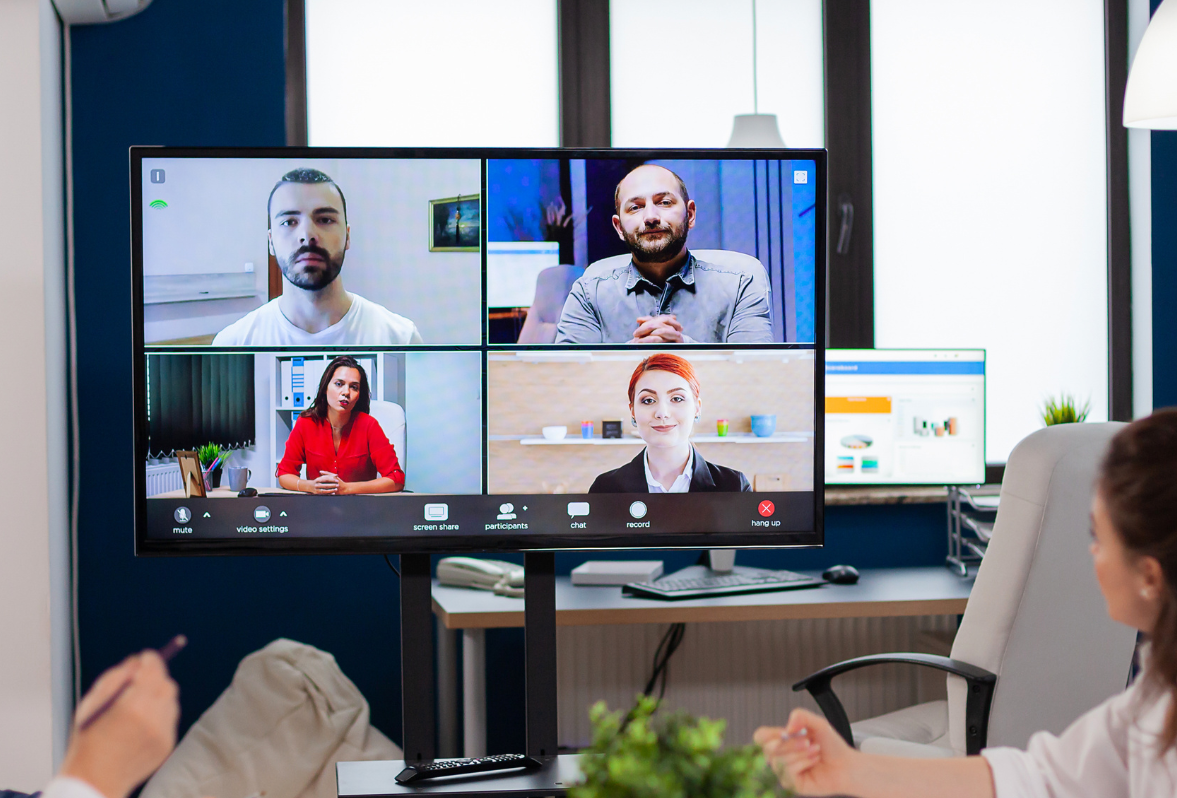 Video conference on a monitor: four people in boxes, a person in a chair, looking at the screen.