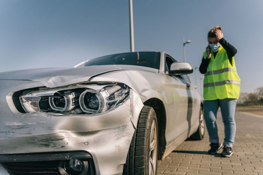 Een vrouw met een masker staat naast een beschadigde auto.