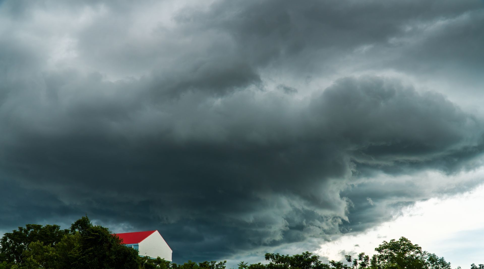 Dark, dramatic storm clouds loom over trees and a building with a bright red roof.