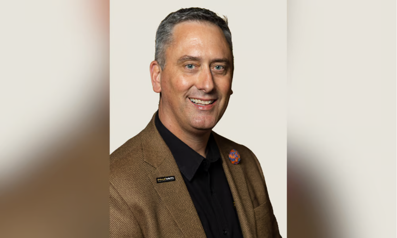 Portrait of a smiling man in a brown blazer and black shirt against a neutral background