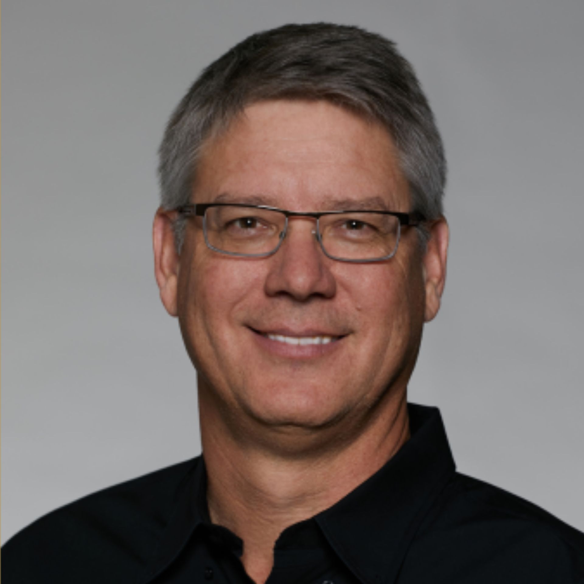A man with gray hair and glasses smiles, wearing a black shirt, against a gray background.