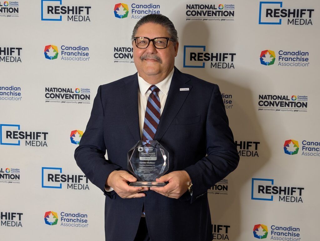 Man holding an award in front of a branded event backdrop, wearing a dark suit and smiling.