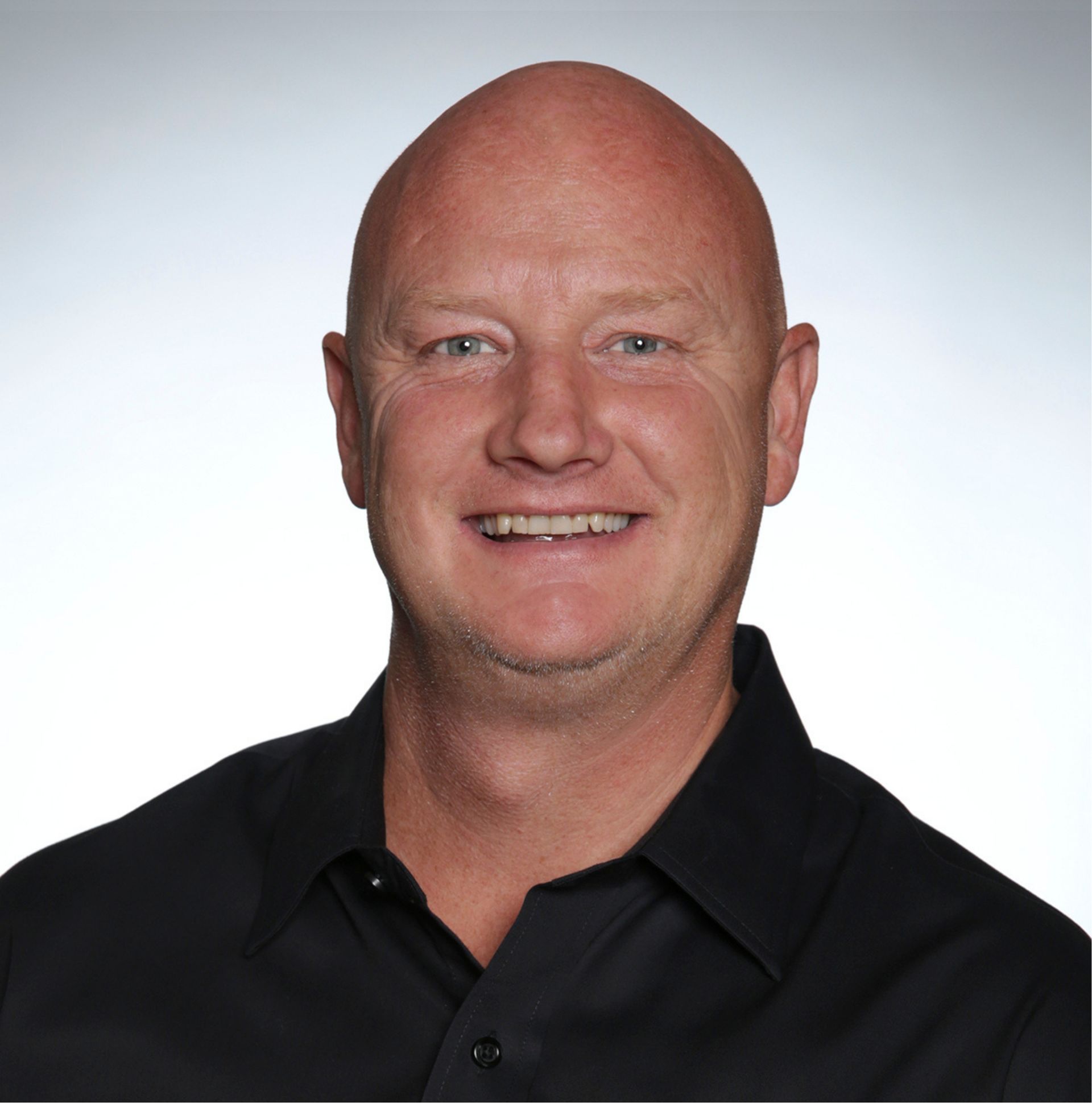 Bald man with fair skin, wearing a black shirt, smiles at the camera against a gradient blue backdrop.