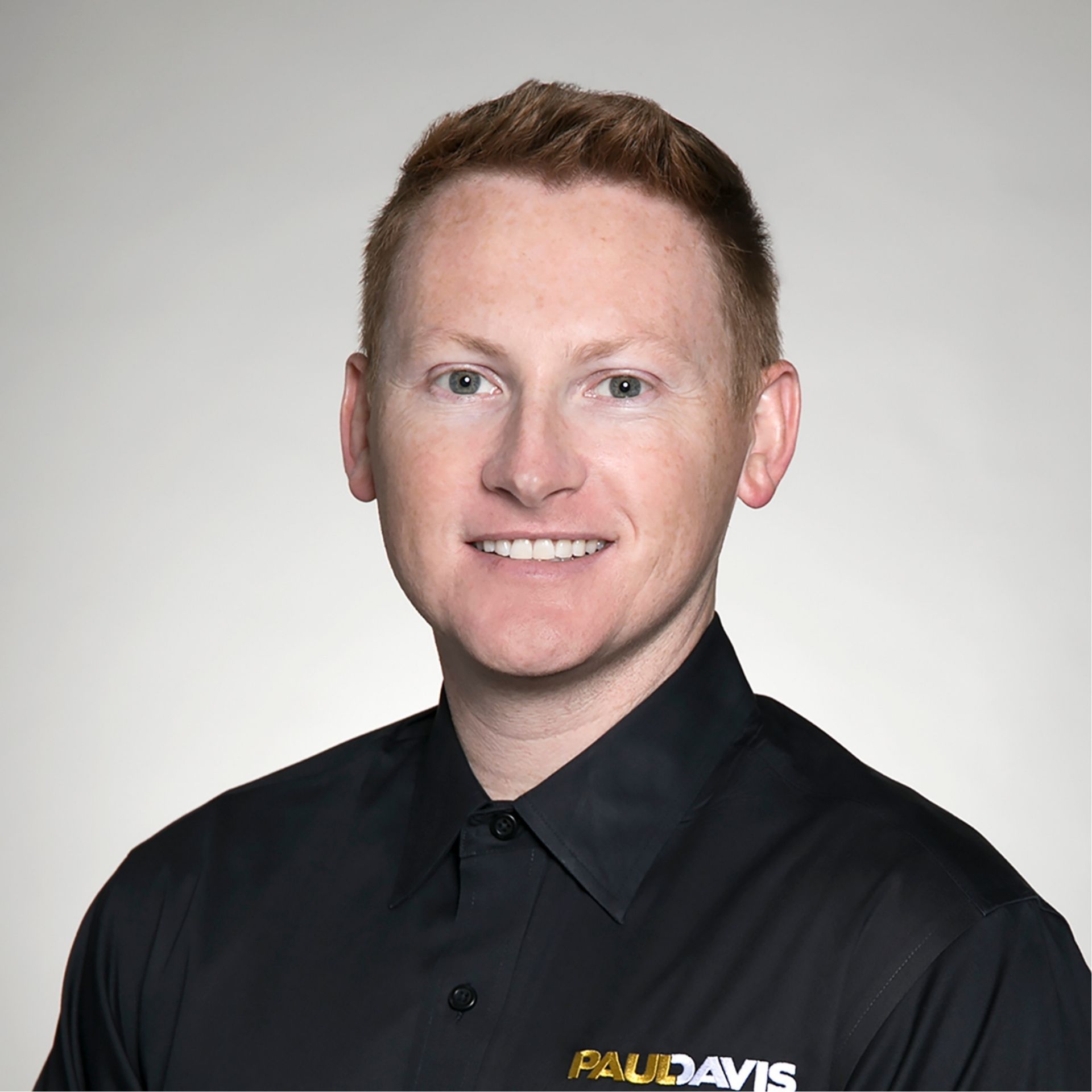 Portrait of a smiling person in a black collared shirt against a gray background, with a logo on the chest