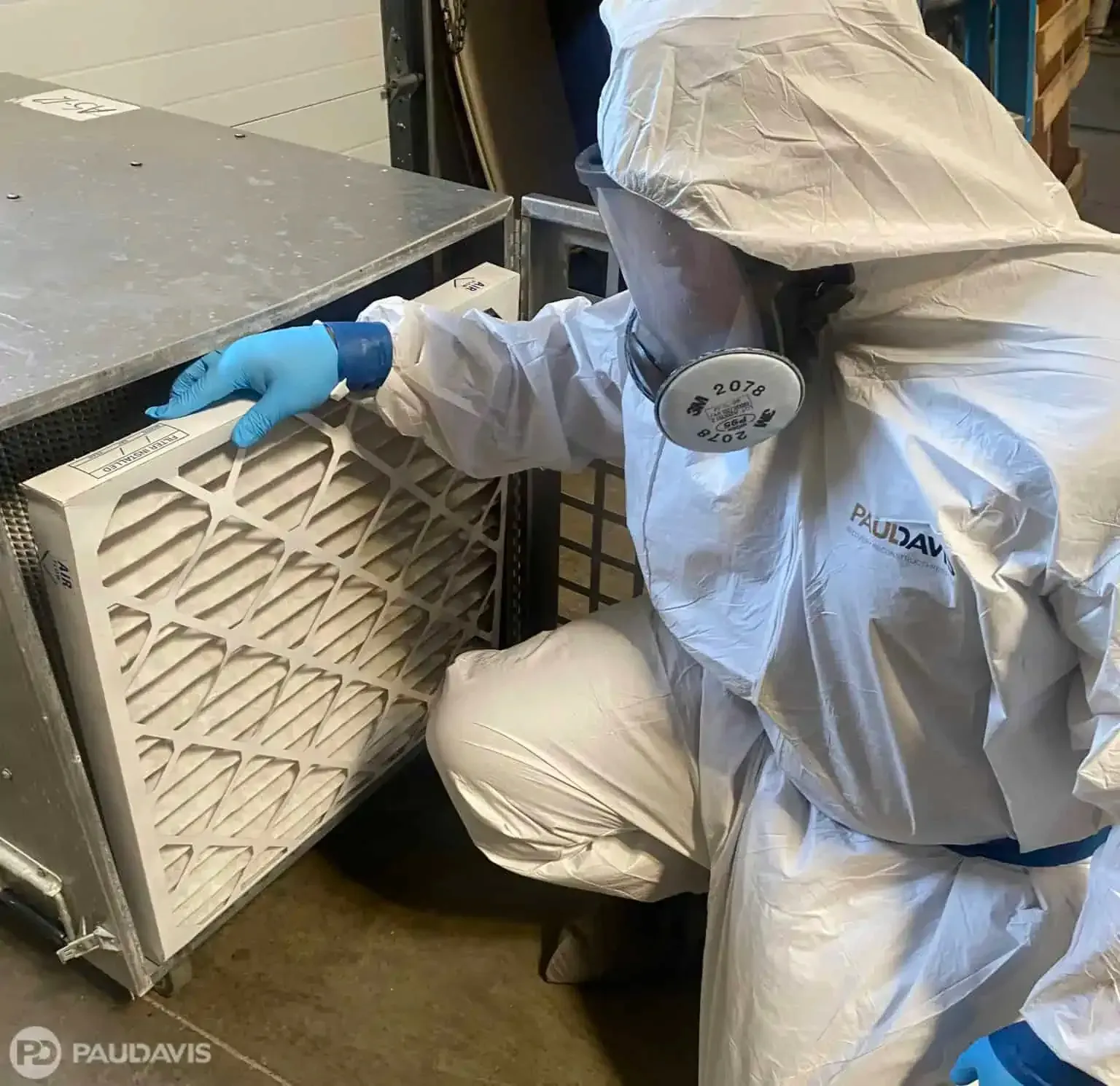 Person in hazmat suit, blue gloves, replacing air filter in industrial setting.