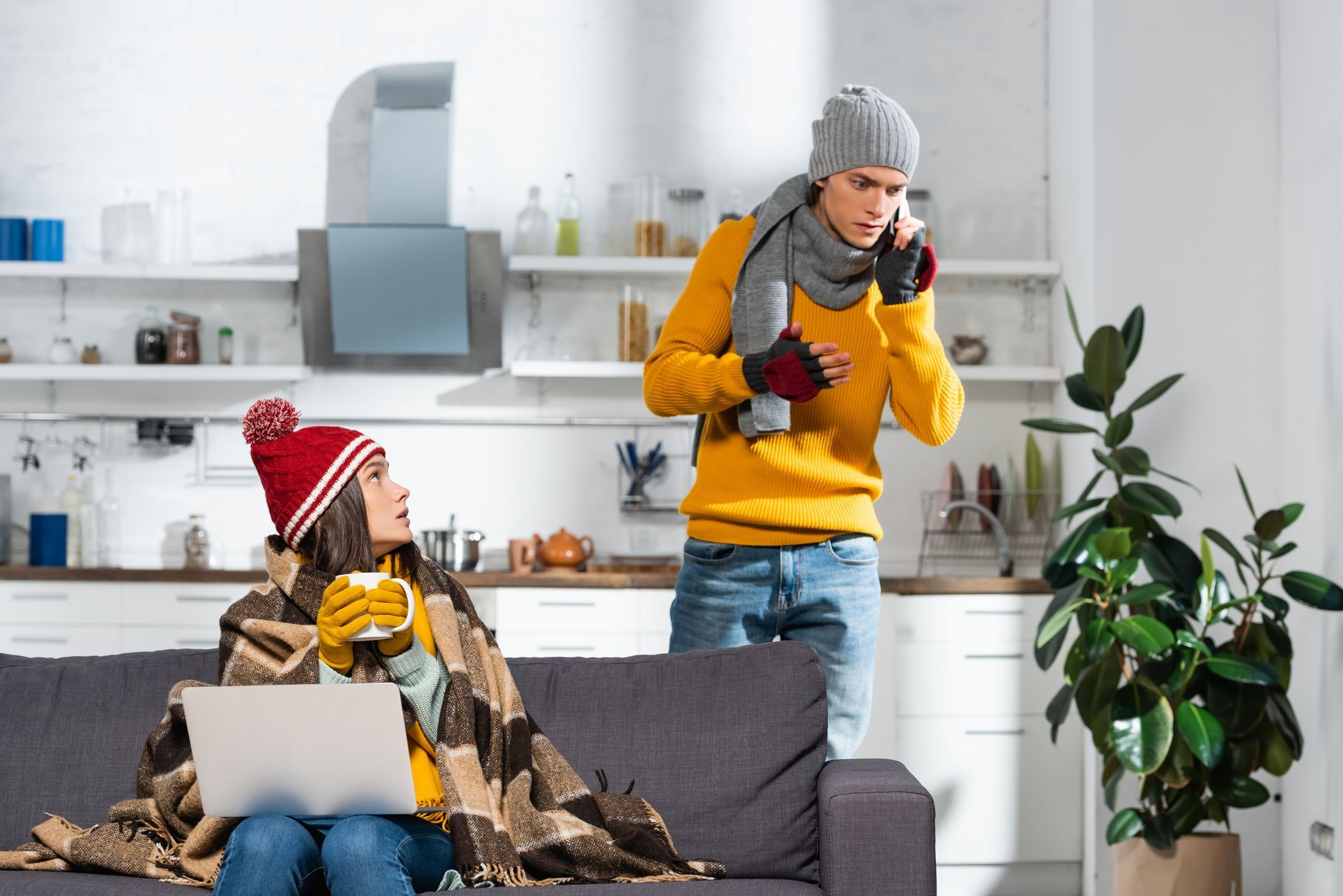 Two people in winter clothes at home, one on a couch with a laptop, one standing by the kitchen counter.