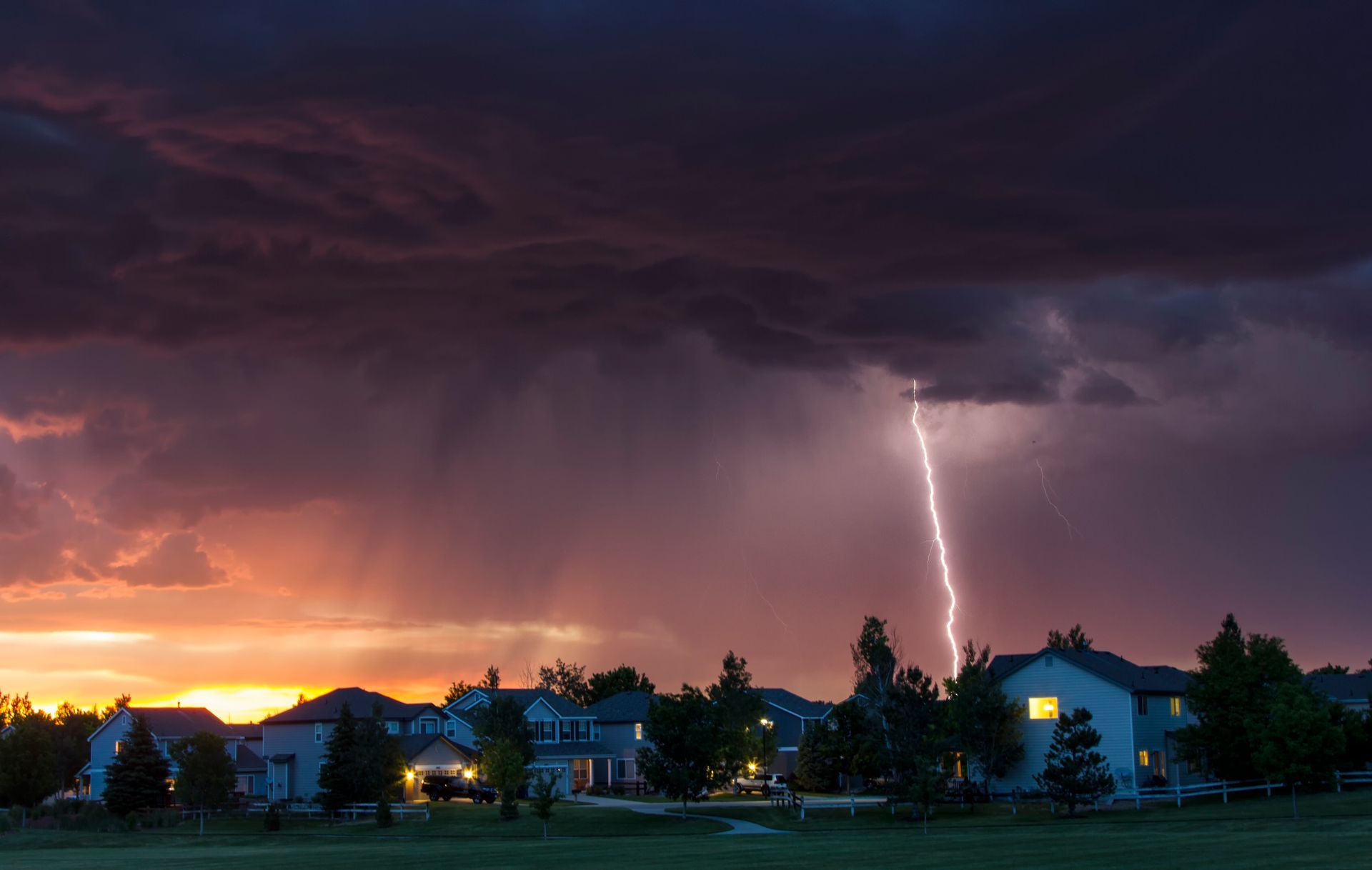 Lightning over a suburban neighborhood at dusk with dark storm clouds and orange sunset glow