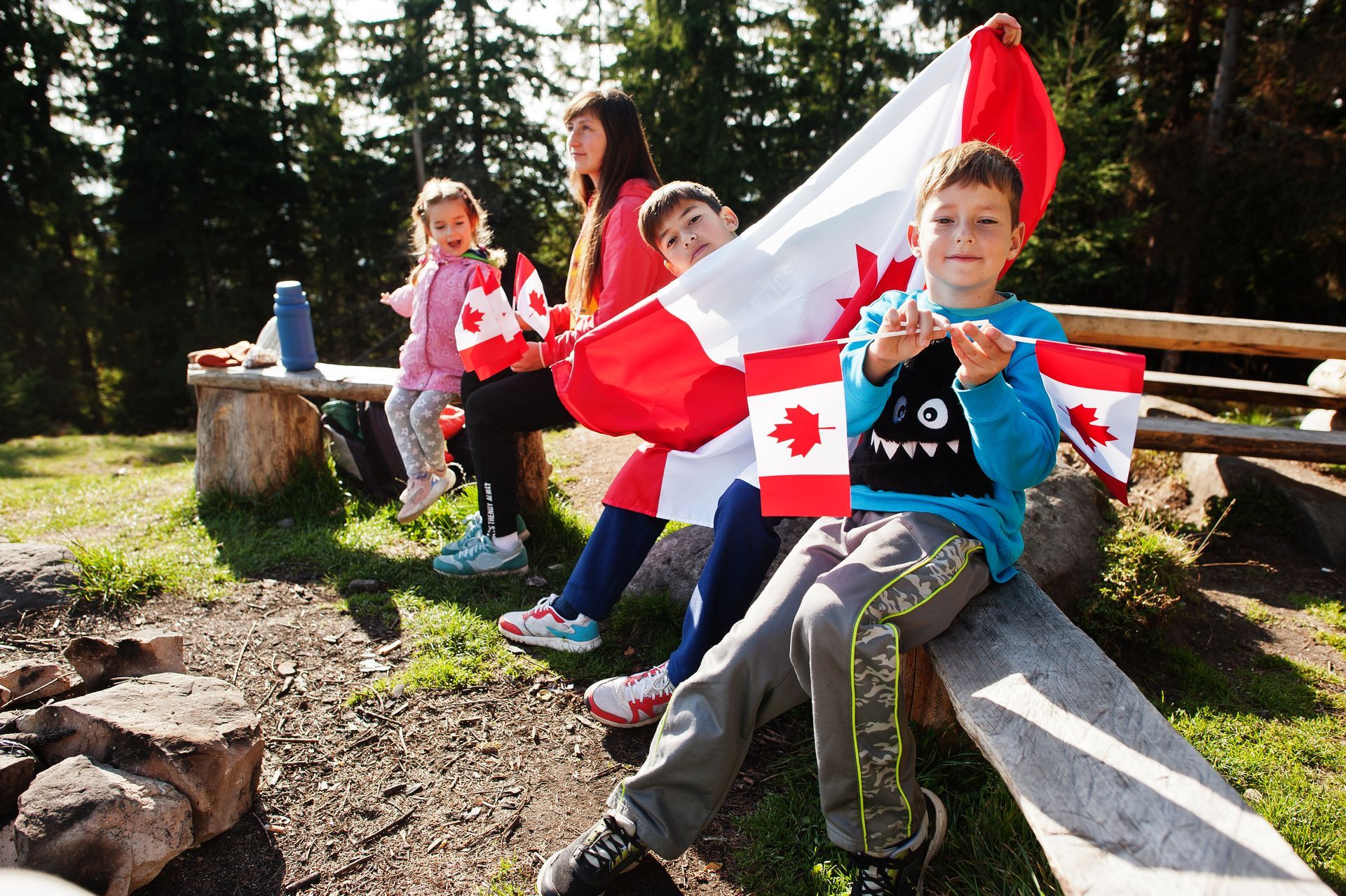 Children sitting on a log bench outdoors, holding a red-and-white Canadian flag in a sunny forest setting