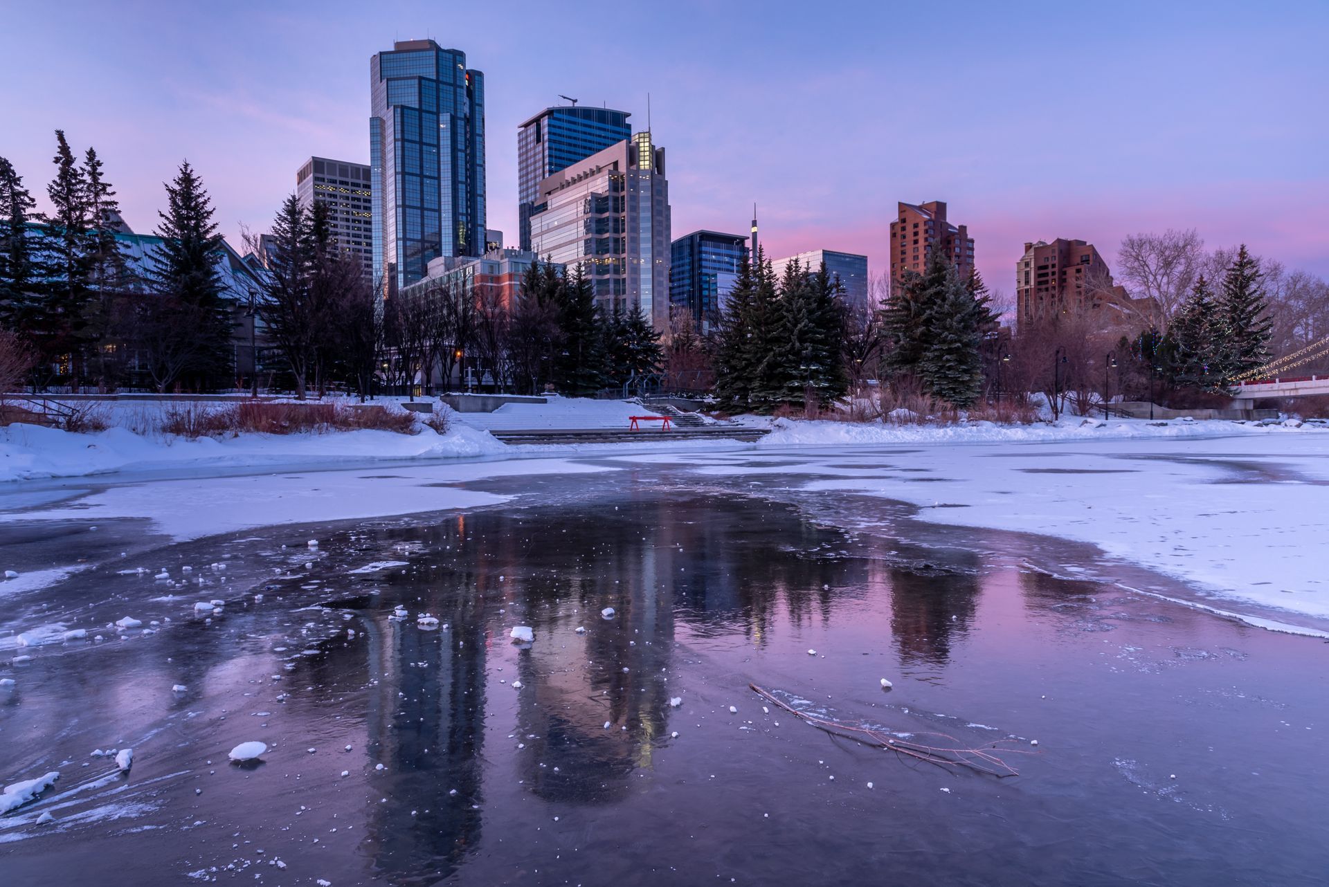 City skyline reflected in a partially frozen river at dusk, with snow and trees in the foreground.