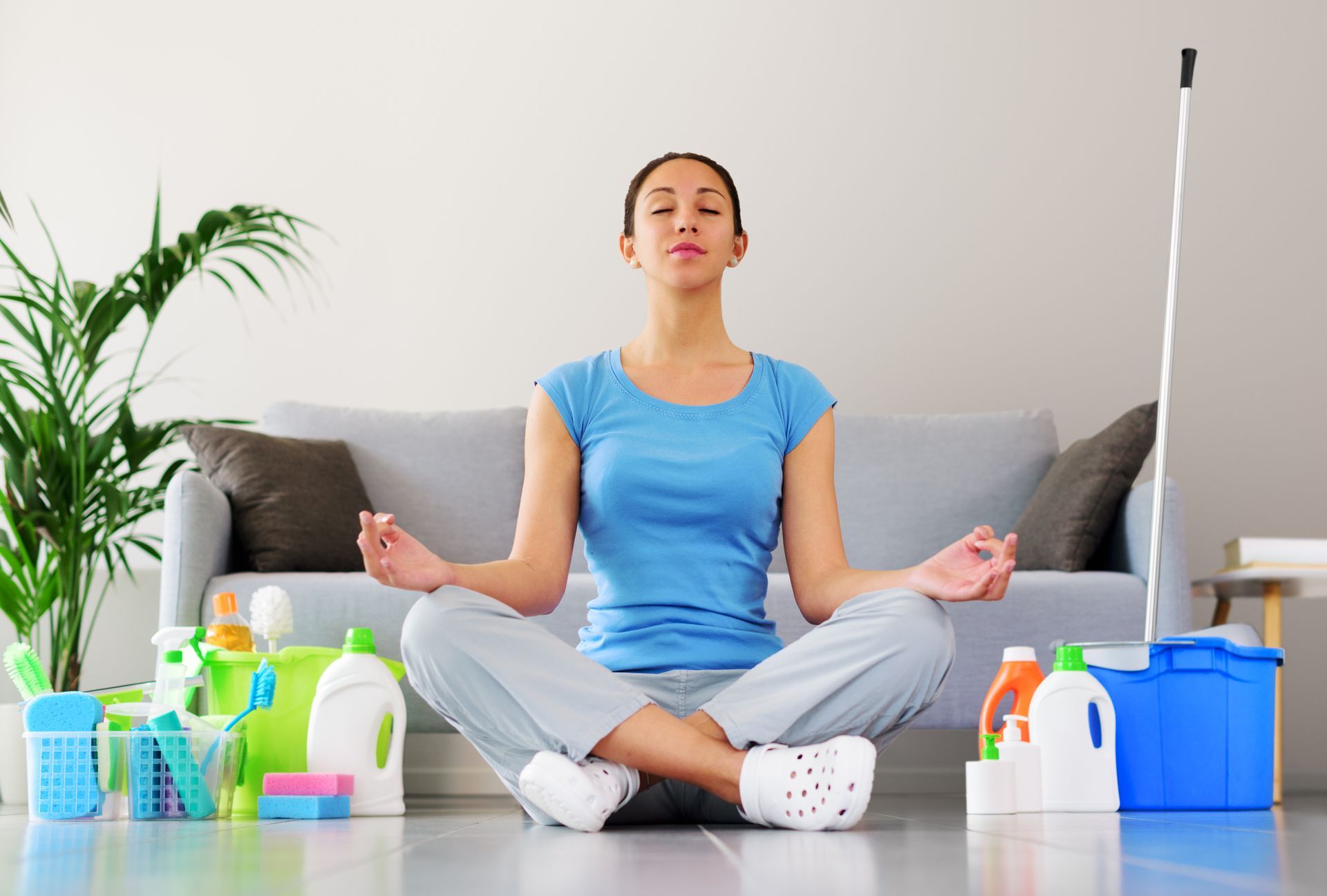 Person meditating on the floor in a bright living room with cleaning supplies nearby