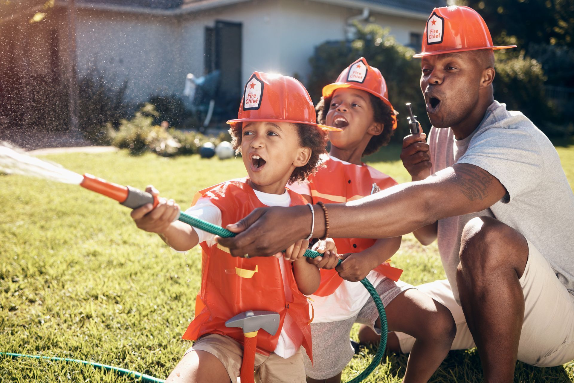 Family in red hard hats holding a garden hose, reacting to water spray on a sunny lawn.