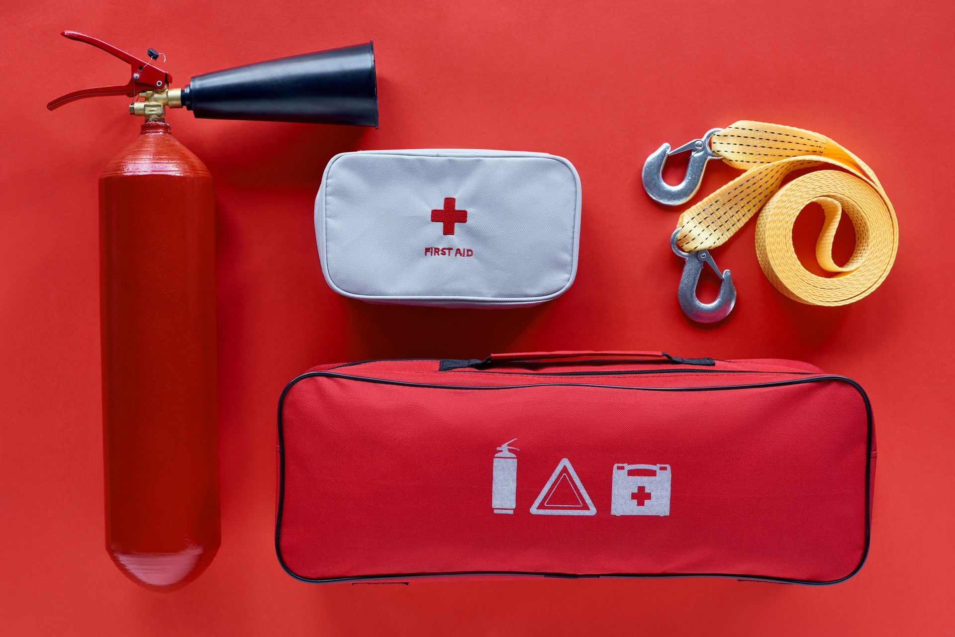 Emergency kit with fire extinguisher, first aid bag, and tow strap on red background.