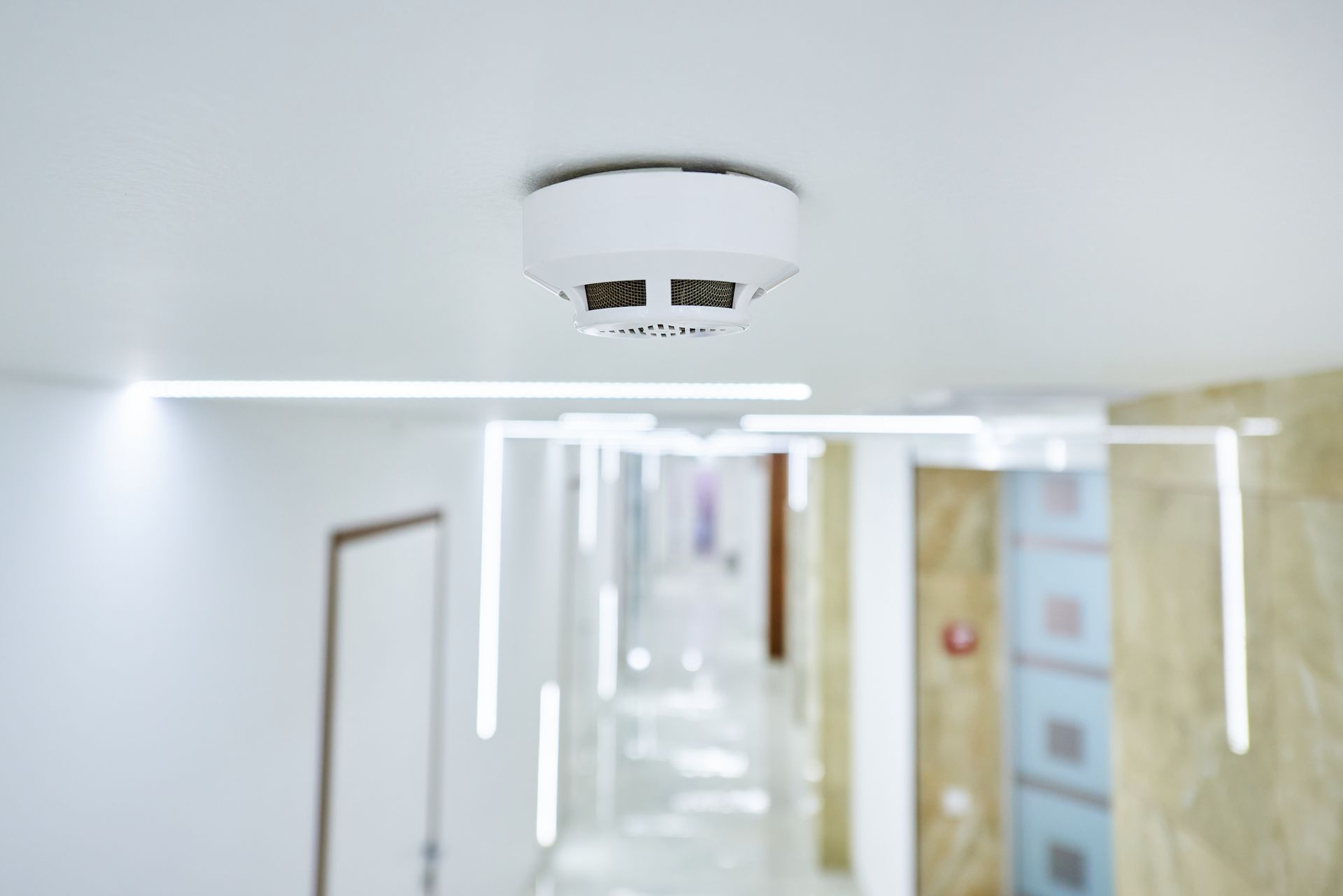 White hallway ceiling with a smoke detector and bright lights reflecting on a polished corridor.