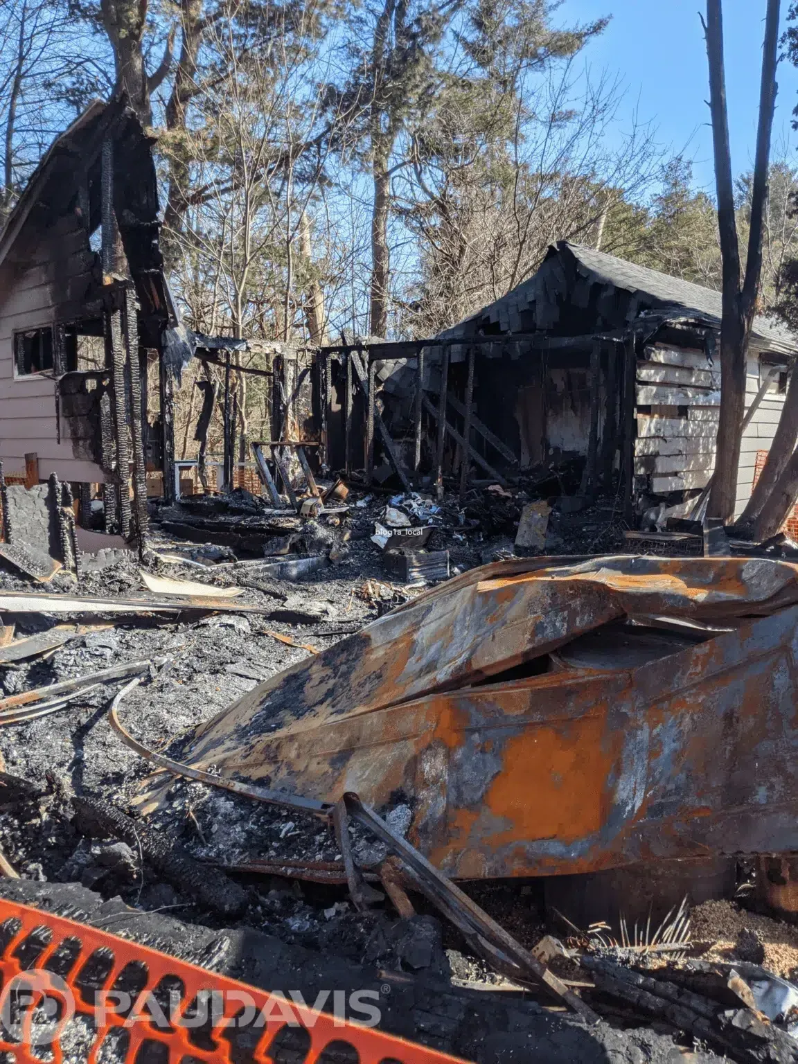 Charred remains of a home after a fire. Burnt wood and debris dominate the scene with caution tape in the foreground.