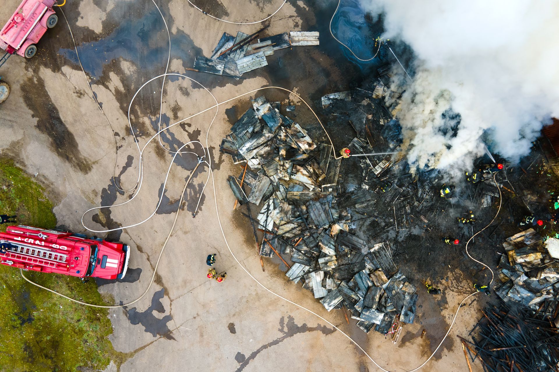 Aerial view of firefighters extinguishing a large structure fire, with smoke, hoses, and debris visible.