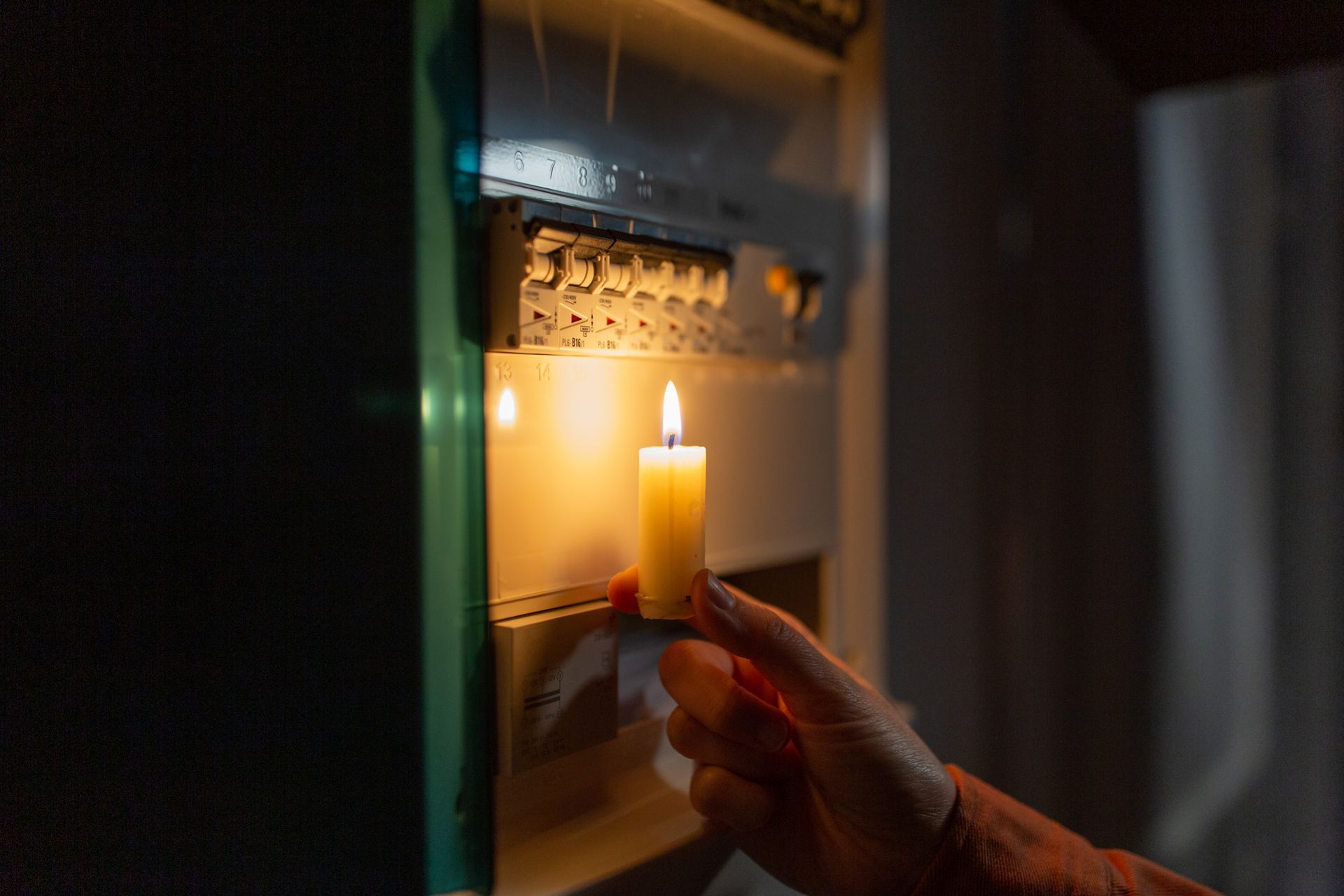 Hand holding lit candle near a fuse box in the dark.