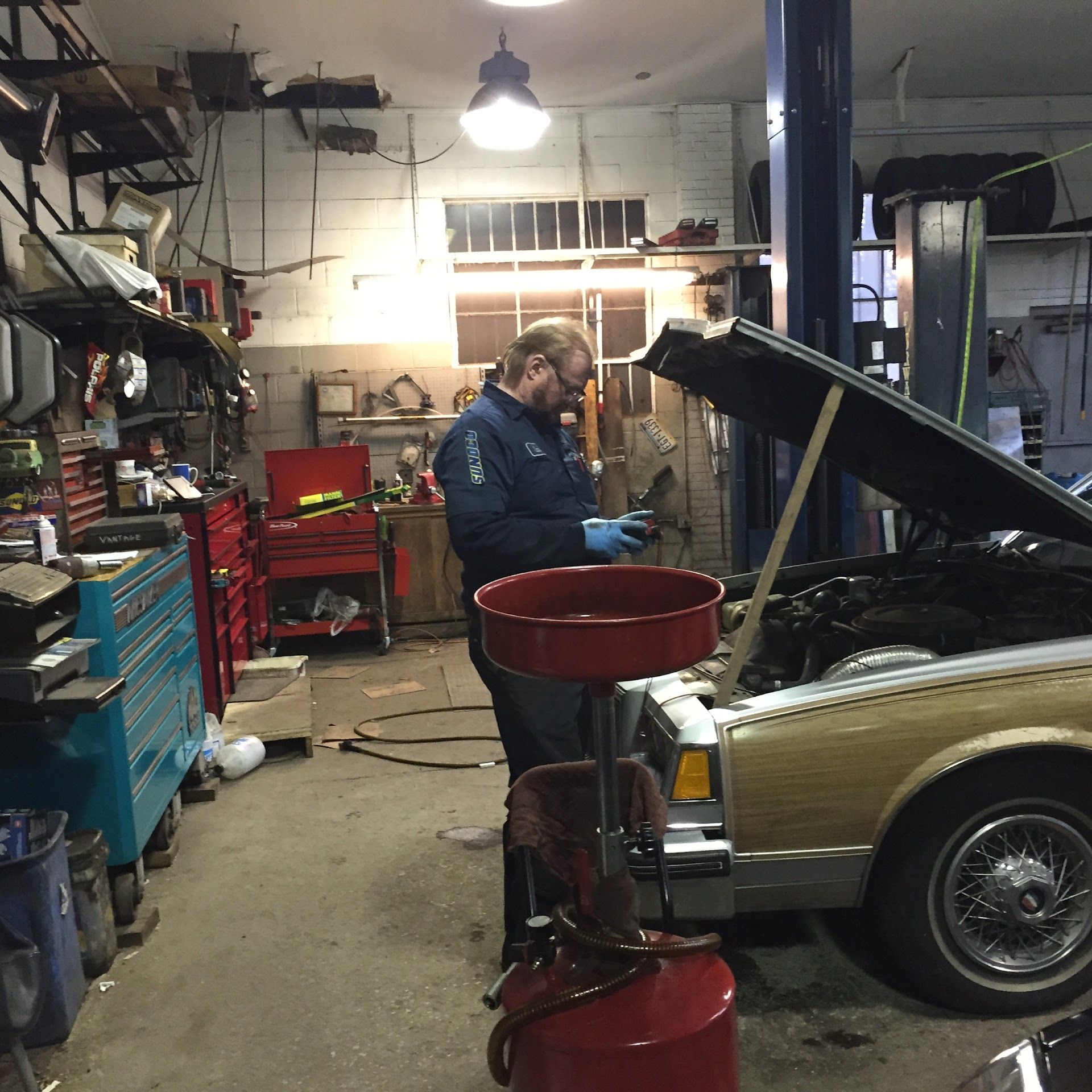 Mechanic in blue coveralls working on car in a cluttered garage, with oil drain pan in place. | Aberdeen Automotive 