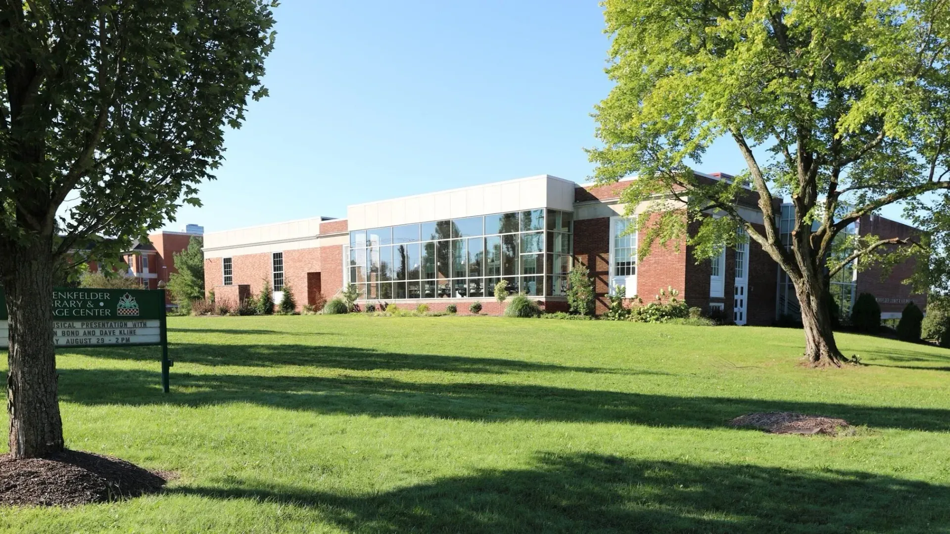 Brick building with large windows on a sunny lawn, framed by trees and a sign in the foreground