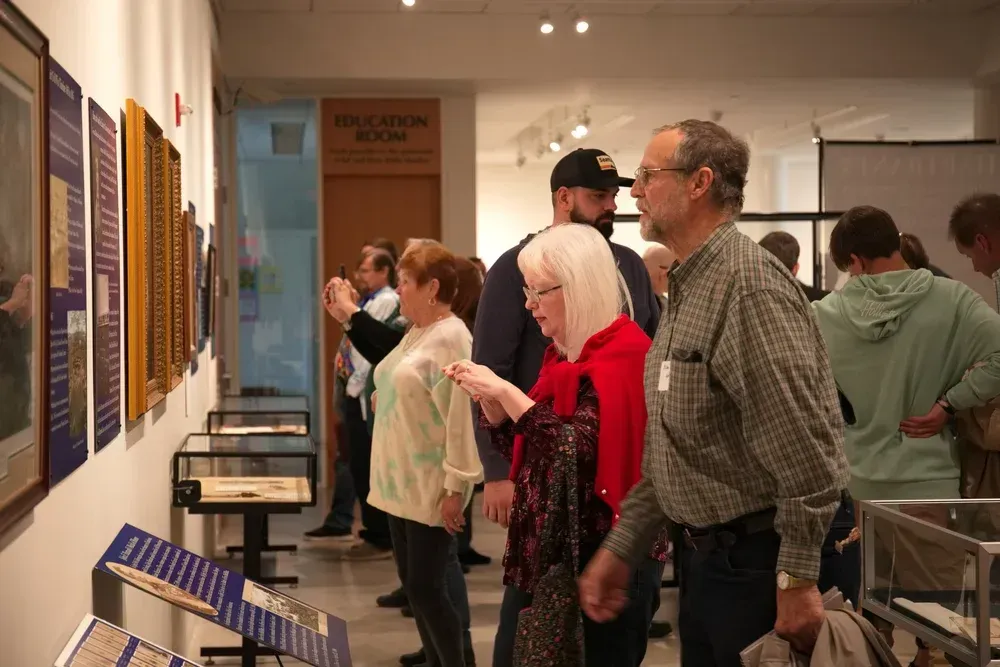 People viewing wall-mounted artworks in a gallery, including a woman in red and a man in a plaid shirt