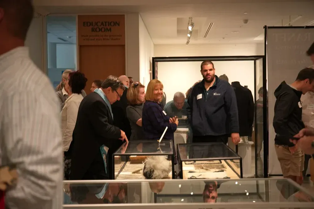People gathered around display cases at a museum exhibit with a guide speaking in the center