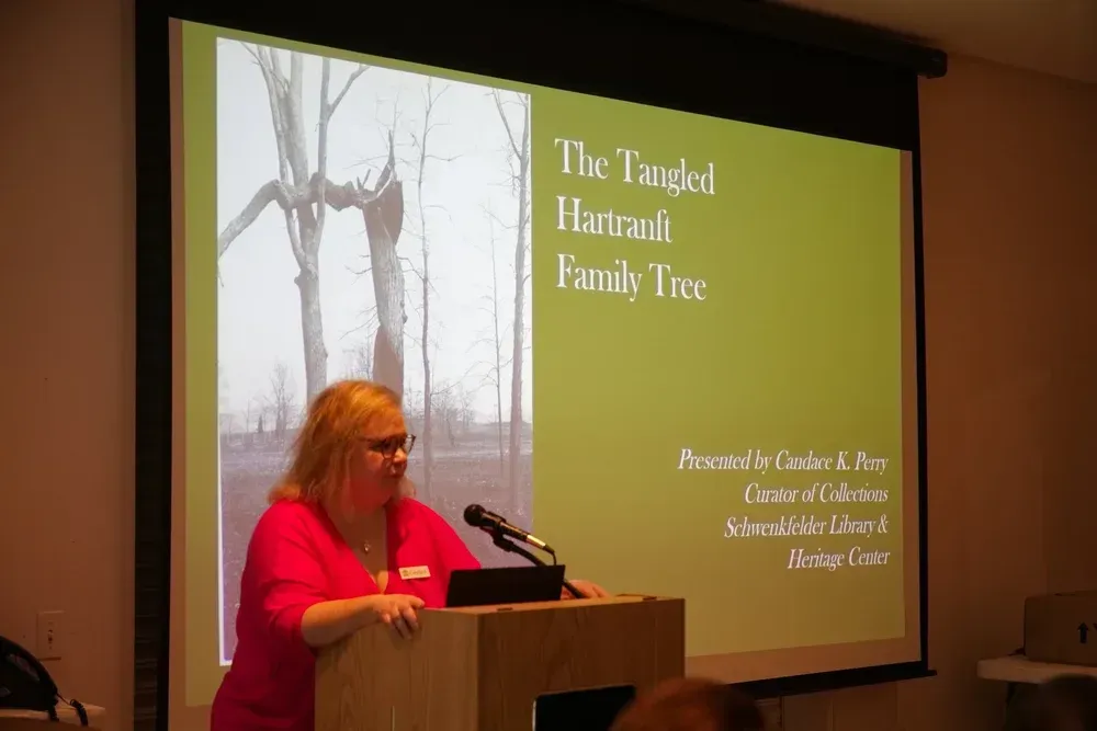 Presenter in pink speaks at podium beside slide titled “The Tangled Hartford Family Tree”