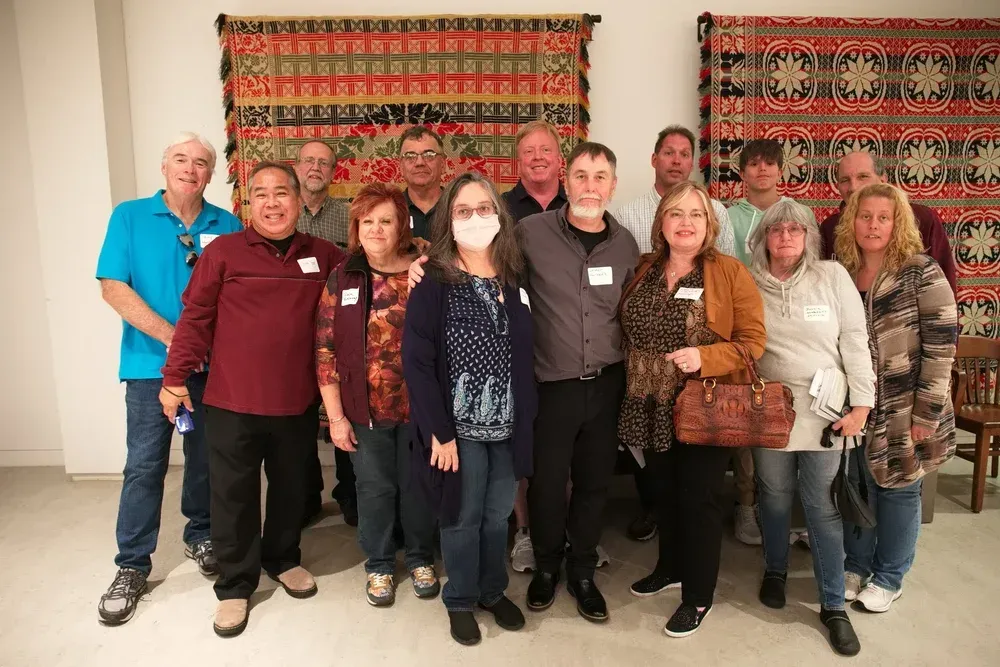Group of people posing indoors in front of a patterned tapestry, wearing name tags and smiling.