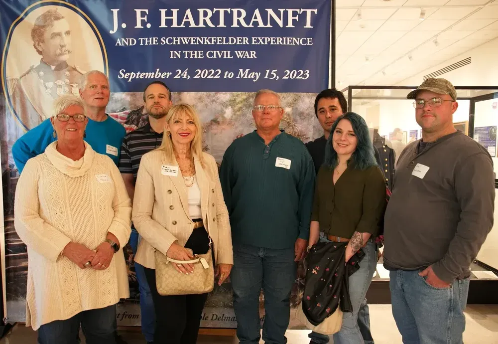 Six people posing indoors in front of a J. F. Hartman exhibit banner.