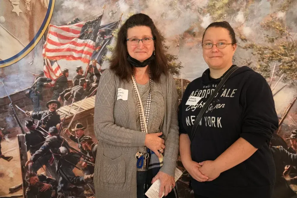 Two women standing before a patriotic Civil War mural, one holding a folded paper and smiling