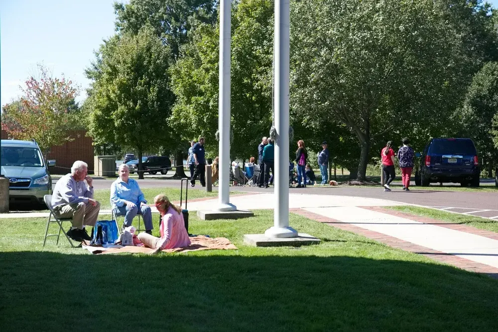 People sitting on grass near a sidewalk and flagpole in a sunny park-like area