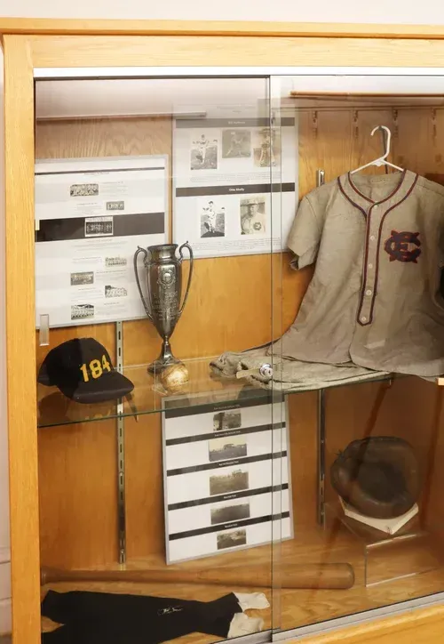Glass display case with a baseball jersey, trophy, cap, and memorabilia.