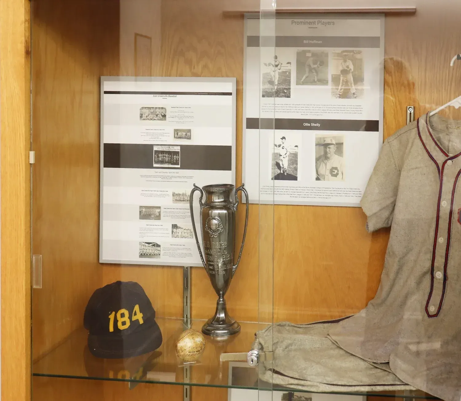 Wooden display case with trophies, a black cap with 184, papers, and a coat on the right